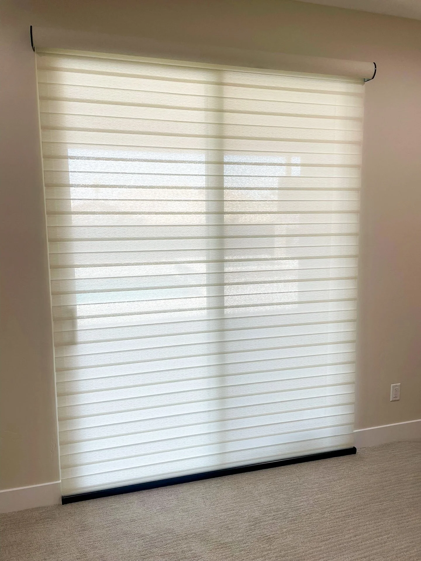 White horizontal window blinds partially open, covering a window in a room with beige walls and carpeted floor.