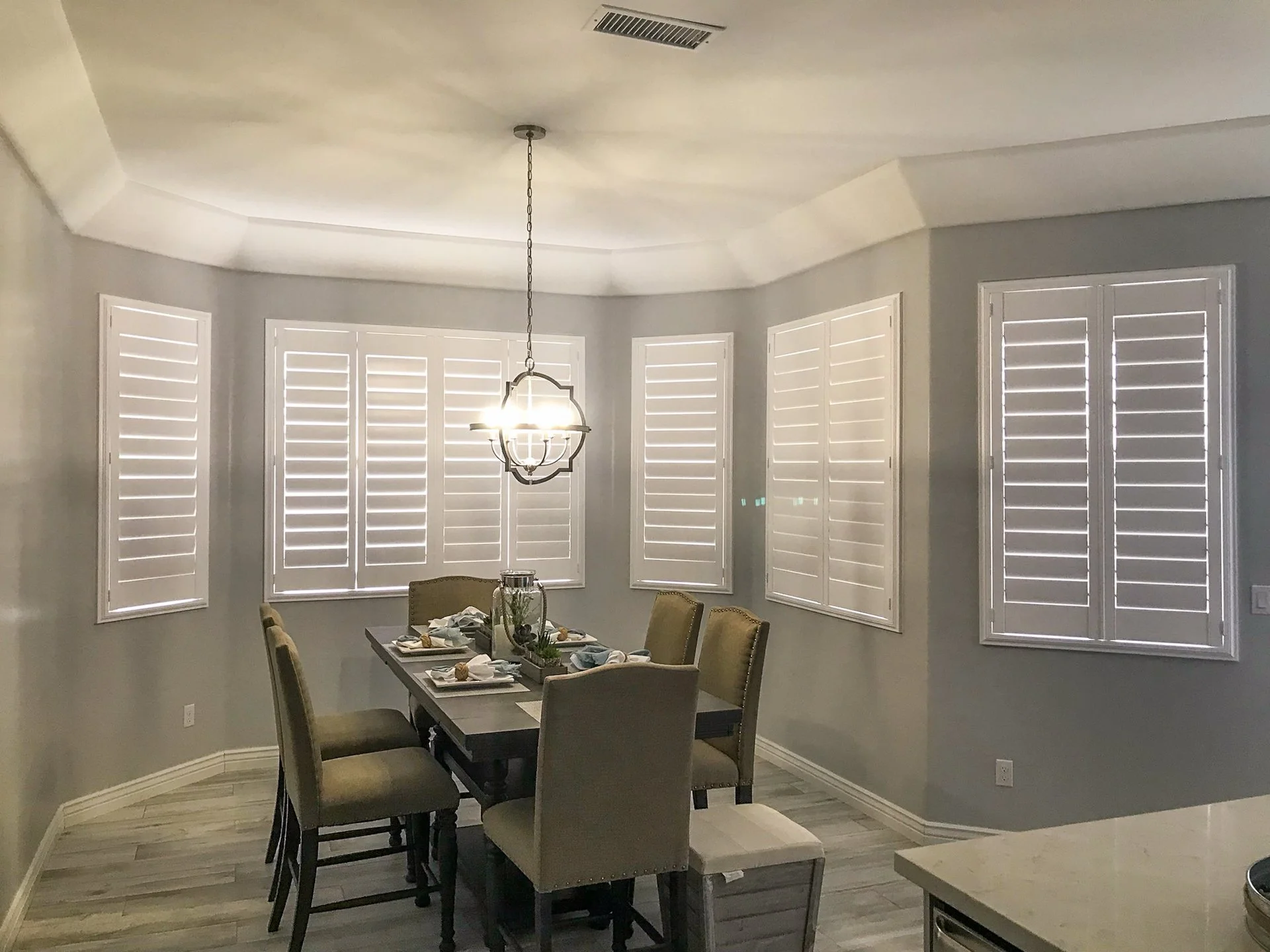 Dining room with round window shutters, a chandelier, and a table set with plates, napkins, and a centerpiece.