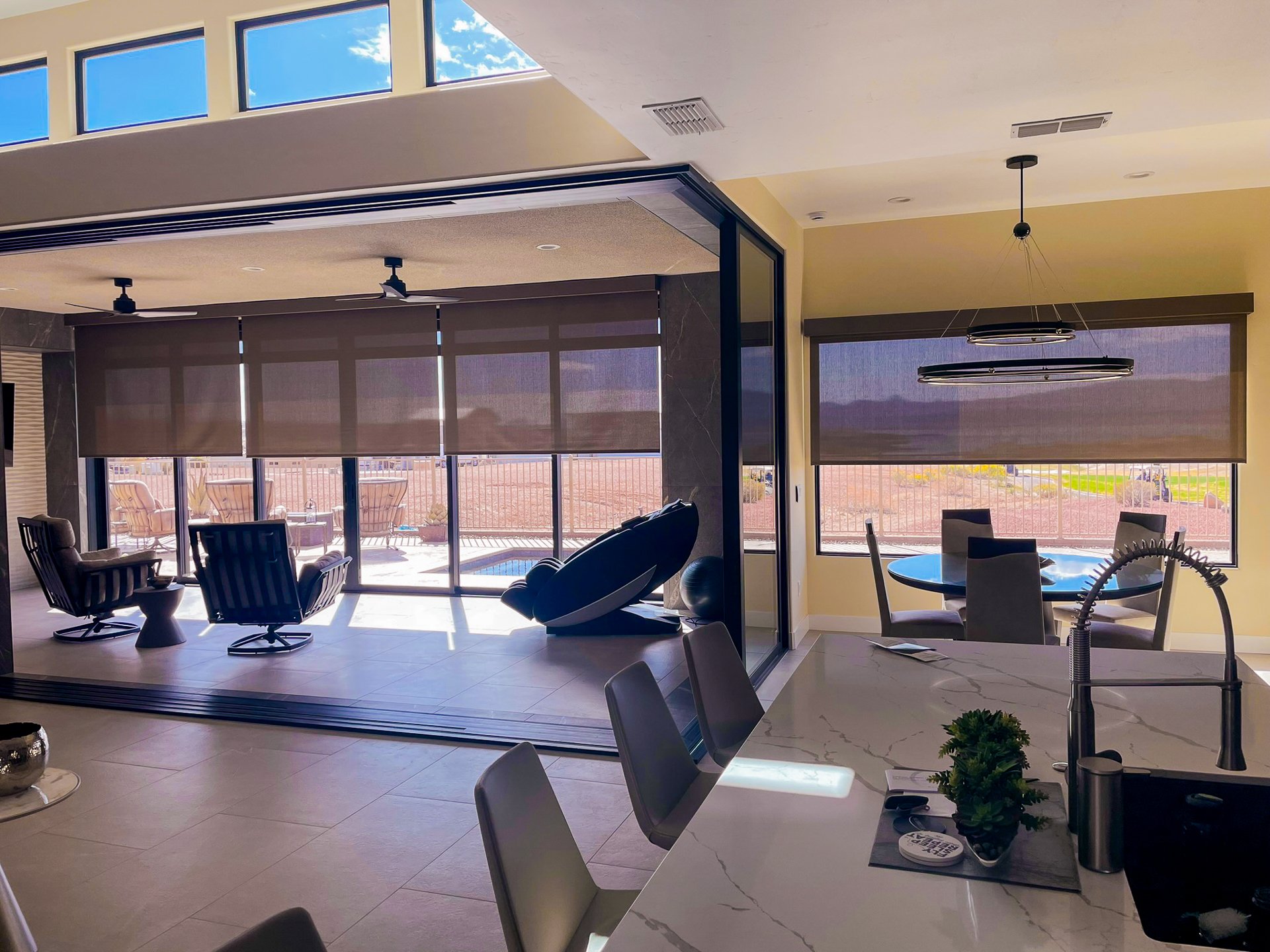 A modern living room with large windows and sliding glass doors leading to an outdoor patio. The room features a seating area with recliners and a rocking chair, a dining area with a round table and chairs, and a kitchen counter with a sink, a small 