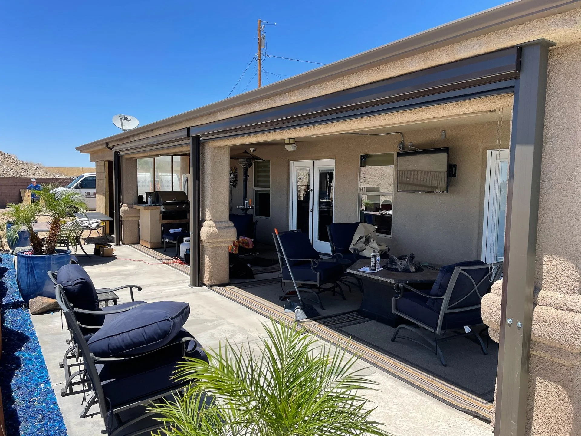 Back patio area with outdoor seating, a small table, a grill, potted plants, and a mounted TV on the house wall under a canopy.