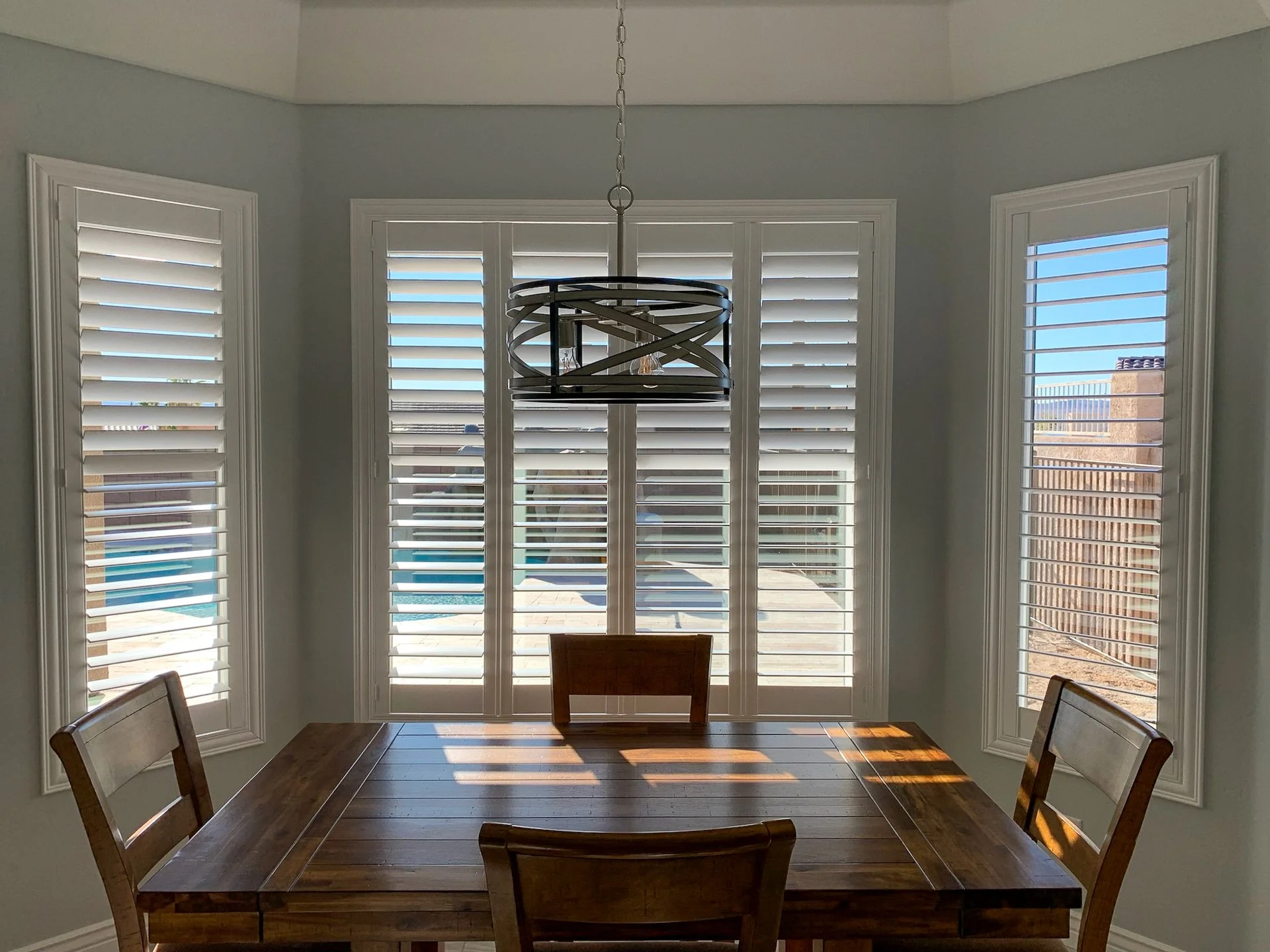 Dining room with a wooden table and chairs, large windows with white shutters, and a modern chandelier hanging from the ceiling.