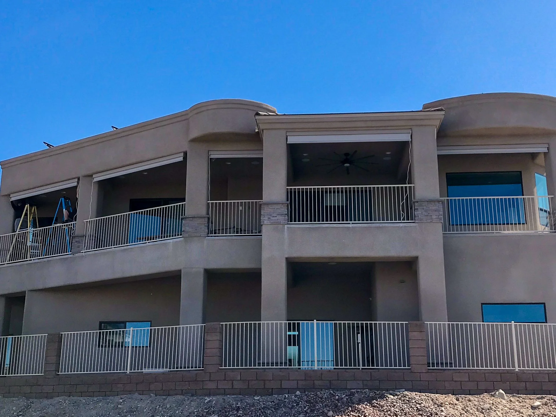 A multi-story residential building with tan stucco exterior, white railing balconies, and large windows, set against a clear blue sky.