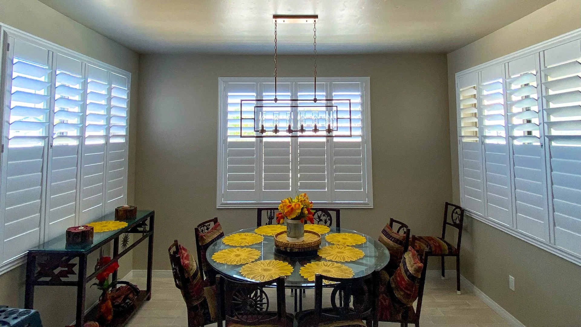 Dining room with a round glass table, yellow sunflower placemats, a floral centerpiece, and six chairs with patterned cushions. White plantation shutters on windows and a modern chandelier overhead.