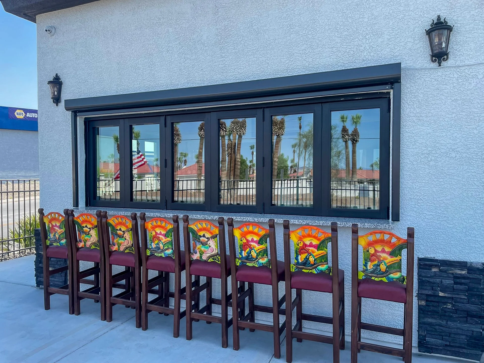 Seven colorful barstools with cartoon parrots and tropical scenery painted on the backrests are lined up in front of a large window with black framing on an outdoor patio. Two black outdoor wall lanterns are mounted on a textured white wall.