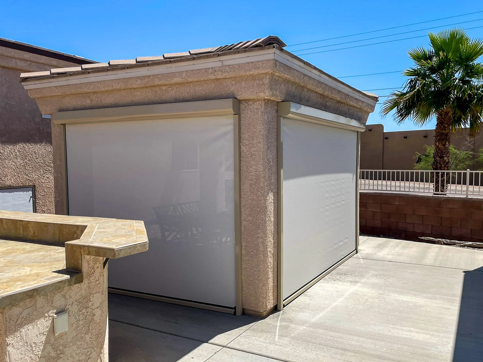 Exterior of a house with two white retractable awnings, a beige stucco wall, and a concrete patio with a curved edge and a beige stone counter. A palm tree is in the background under a bright blue sky.