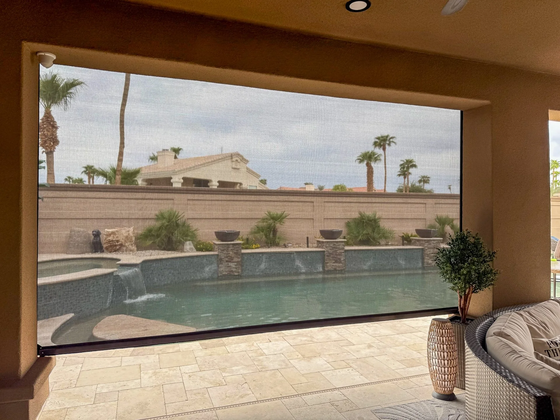 Indoor living space with a large window looking out onto a backyard pool area featuring a waterfall, water bowls, palm trees, a stone wall, and a cloudy sky.