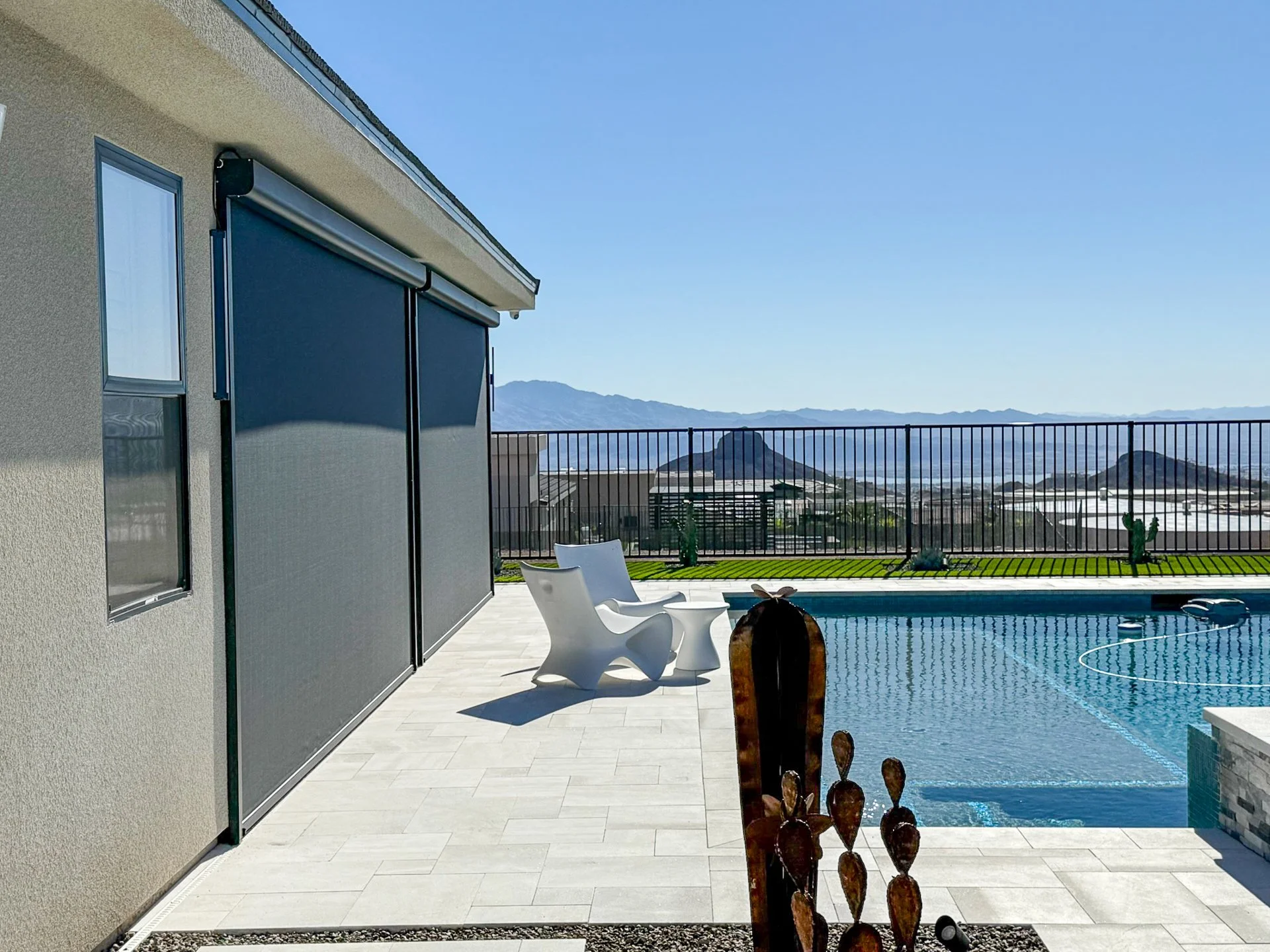 A backyard with a swimming pool, white chairs, a fountain, and mountains in the distance under a clear blue sky.