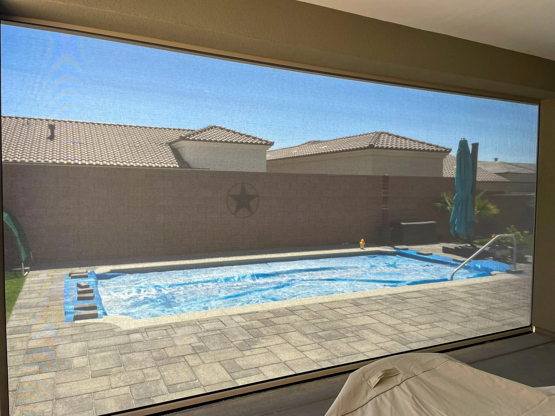 Backyard with a small blue swimming pool, brick patio, and a brick wall with a decorative star. There's a closed blue patio umbrella, some plants, and a blue pool cover.