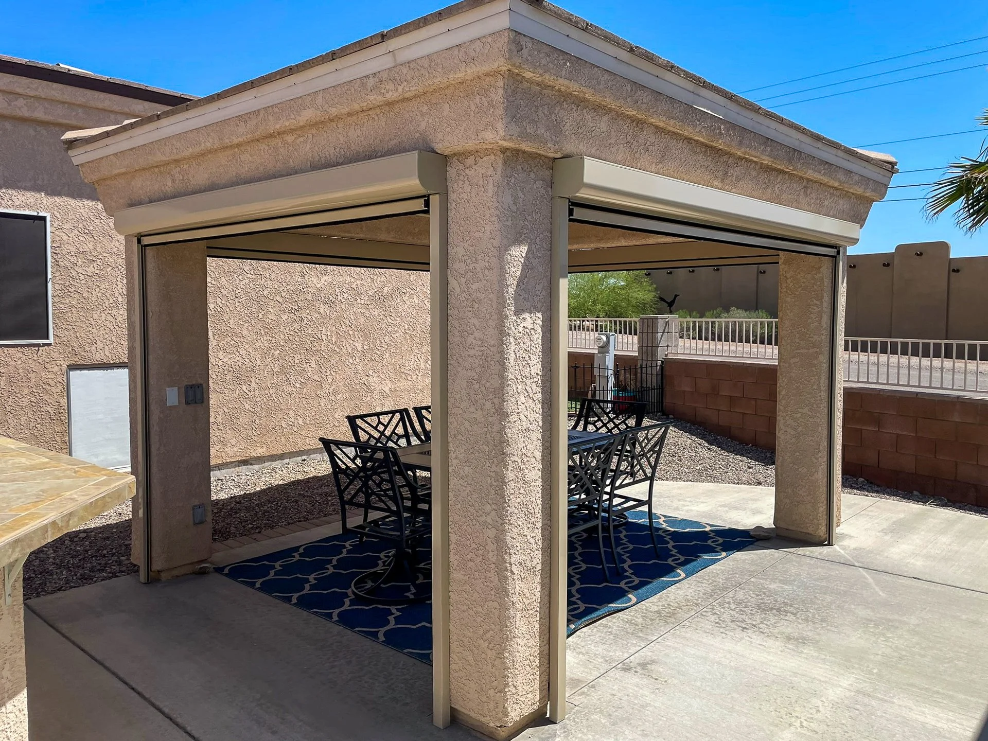 Outdoor patio area with a covered pergola, black metal table and chairs, and a blue geometric rug on a concrete floor, surrounded by a stucco wall and fence, with trees and blue sky in the background.