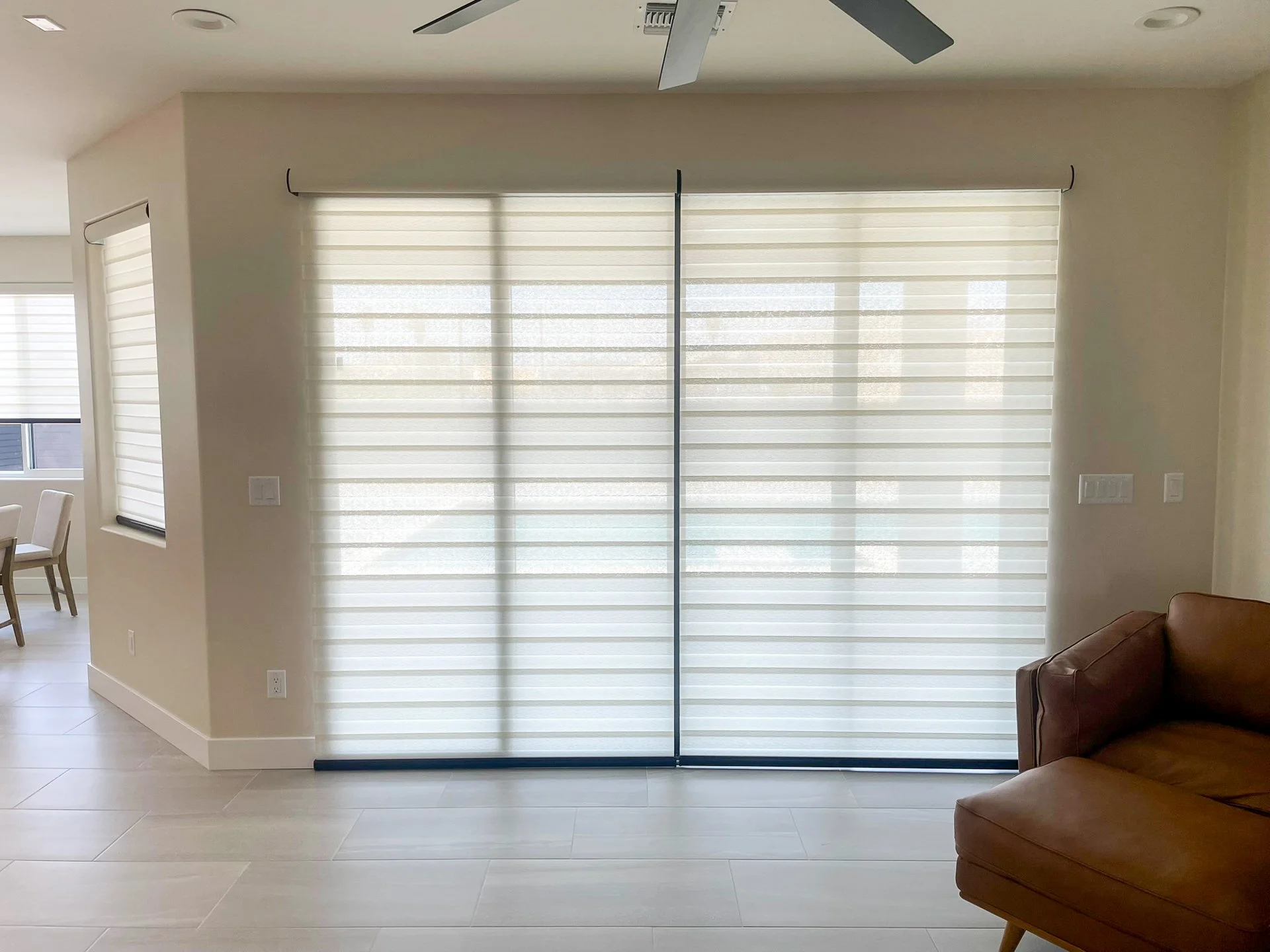 Living room with beige walls, a brown leather couch on the right, and large sliding glass doors with white blinds in the center. A ceiling fan is visible at the top.