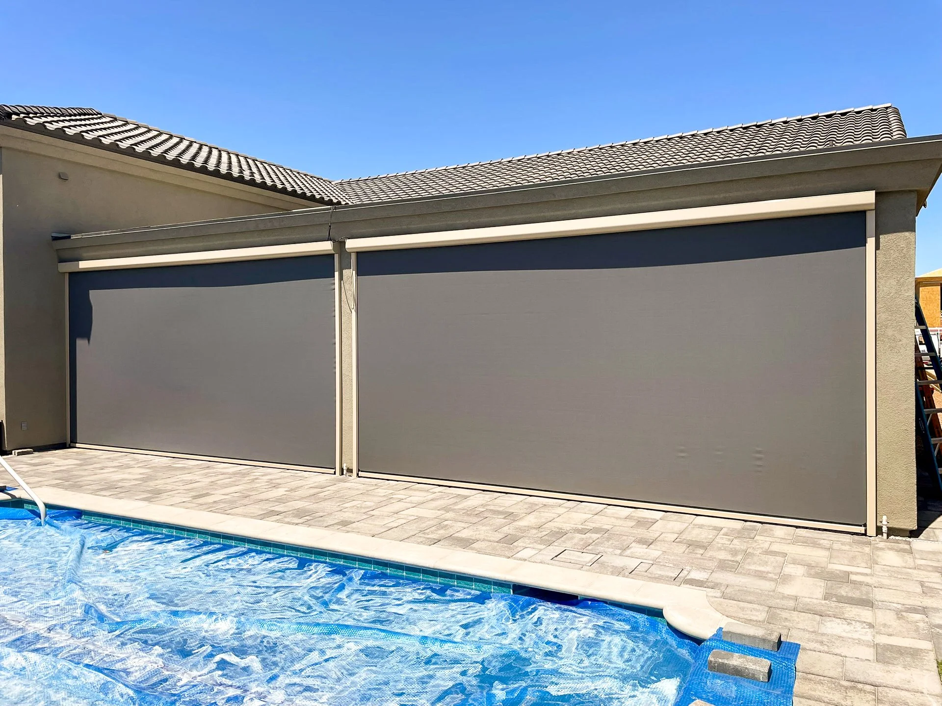 Two closed gray retractable pool enclosures next to a swimming pool filled with water, on a paved patio under a clear blue sky.