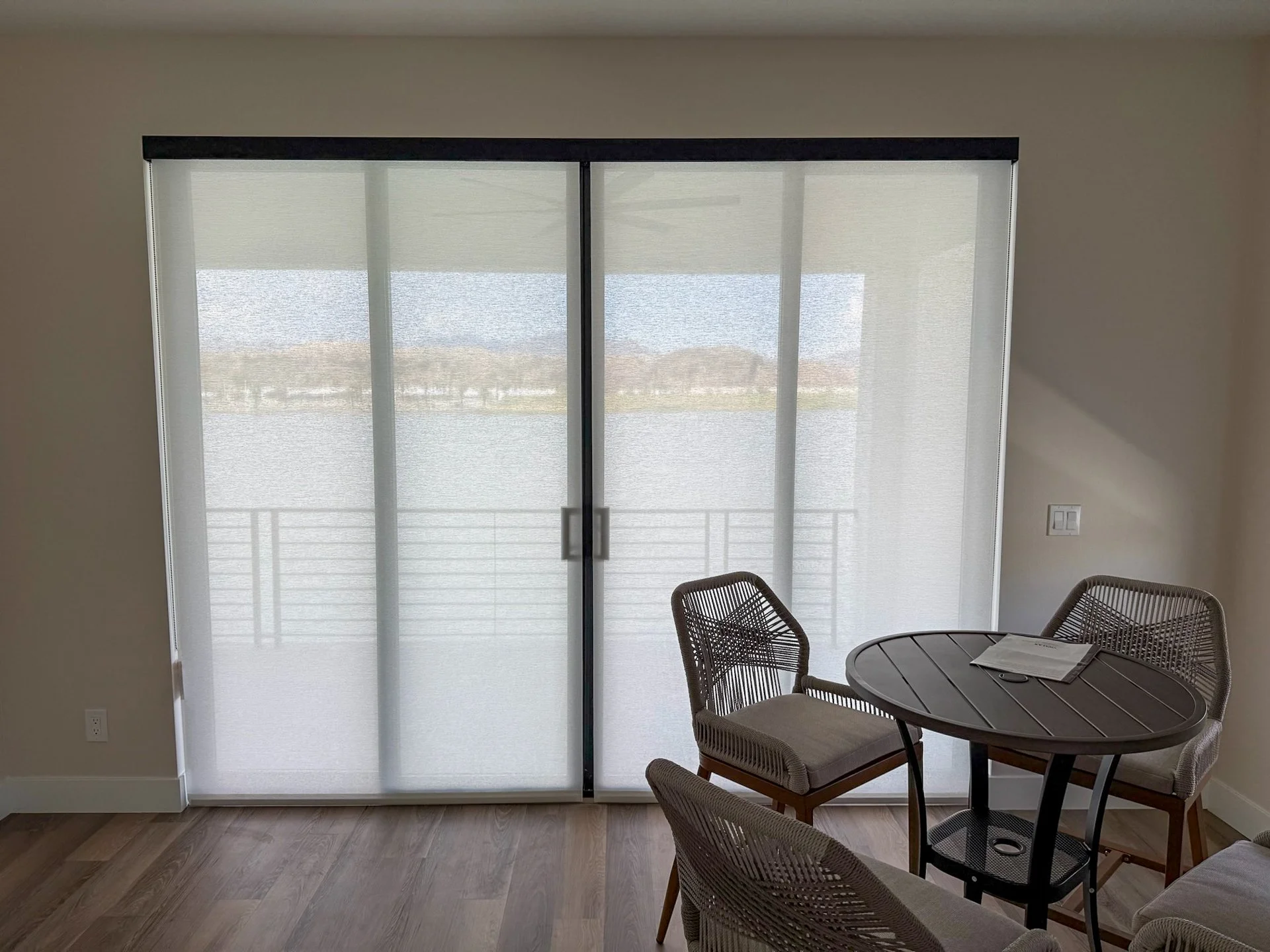 Interior of a room with a sliding glass door covered by a sheer curtain, overlooking a balcony and a body of water outside. There is a small round table with three chairs inside.