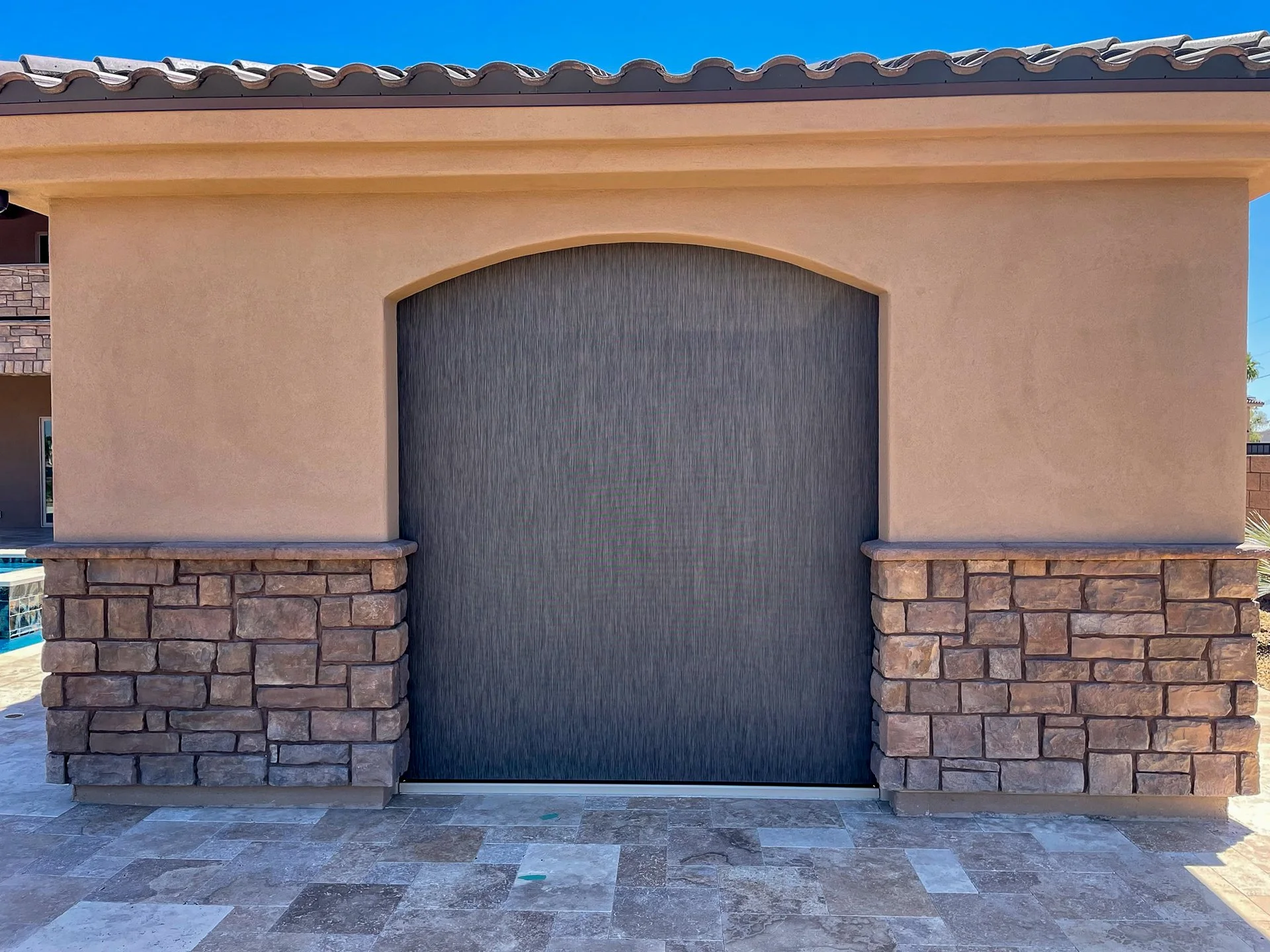 Exterior of a building with a large, arched, dark-colored door, beige stucco walls, and stone accents at the base, with a tile roof and blue sky above.
