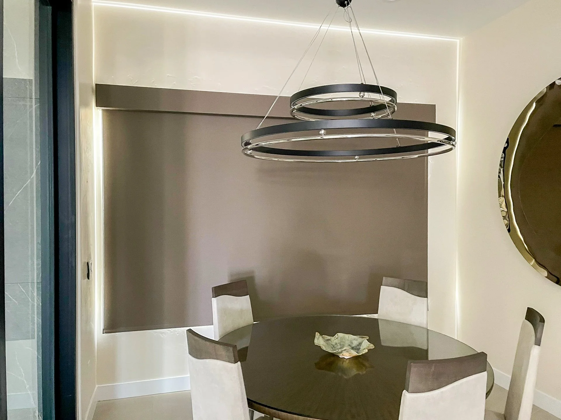 A modern dining area with a round glass-top table, four chairs with light-colored fabric seats, a decorative wooden bowl in the center, a wall with a gray-colored panel and LED lighting, and a large circular hanging light fixture above the table.