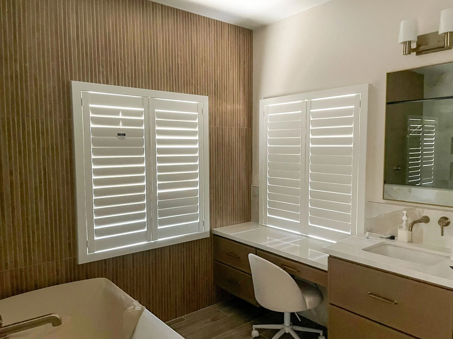 Bedroom with two windows featuring white shutters, wooden dresser with a white chair, bathroom sink with soap dispenser, mirror, and wall-mounted lights.