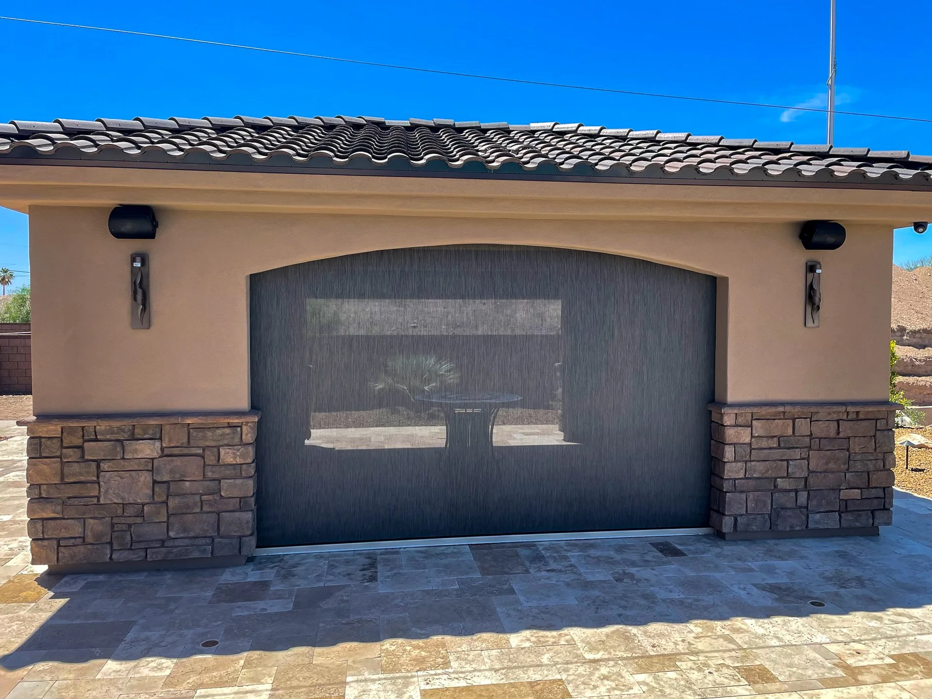 Modern garage door with black mesh screen, stone and stucco exterior, two black wall-mounted lights, on a paved patio under a blue sky.