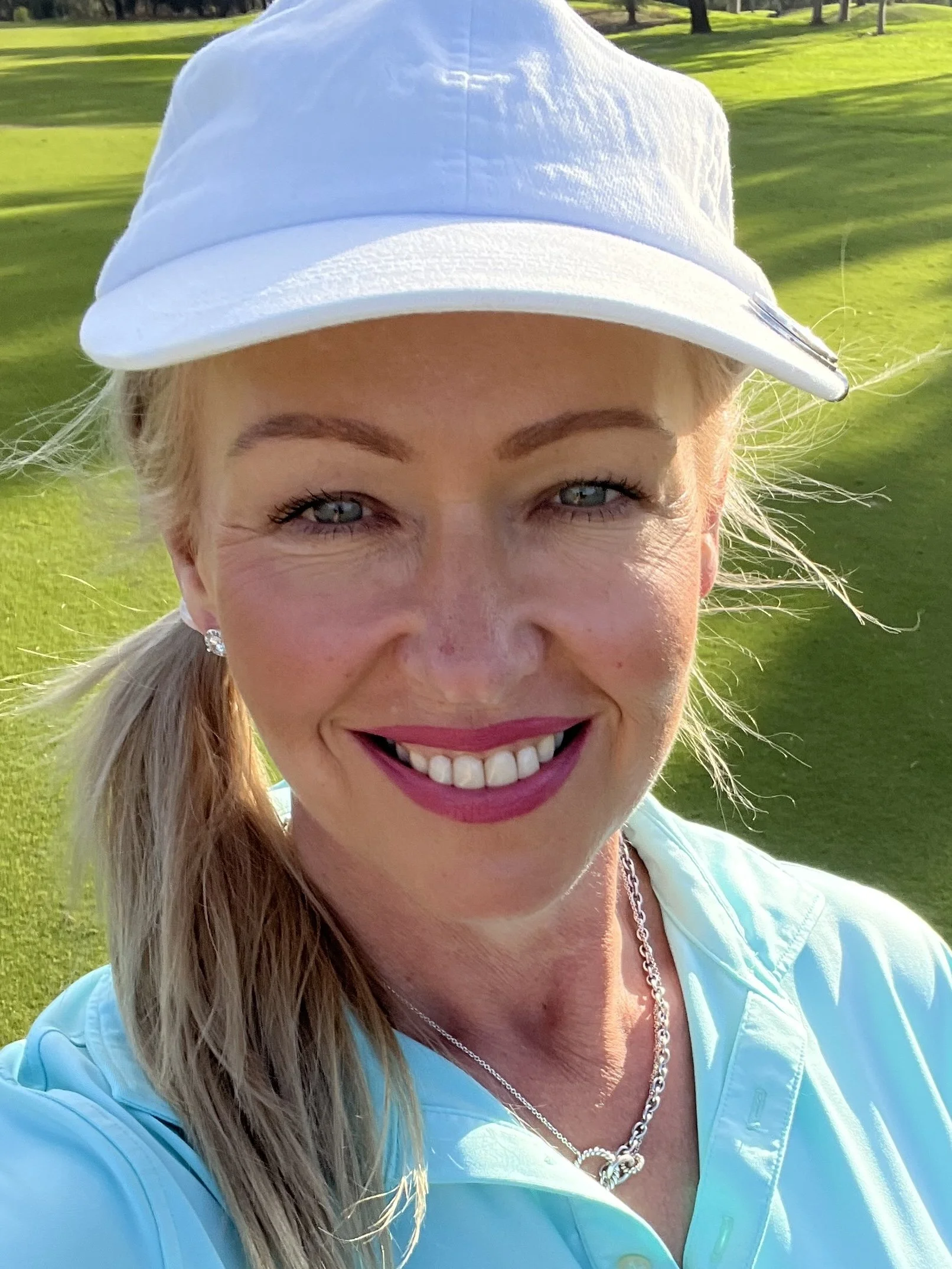 A woman with blonde hair, blue eyes, and wearing pink lipstick, smiling outdoors on a sunny day. She has earrings, a chain necklace, and is wearing a white cap and light blue jacket, with a golf course and trees in the background.