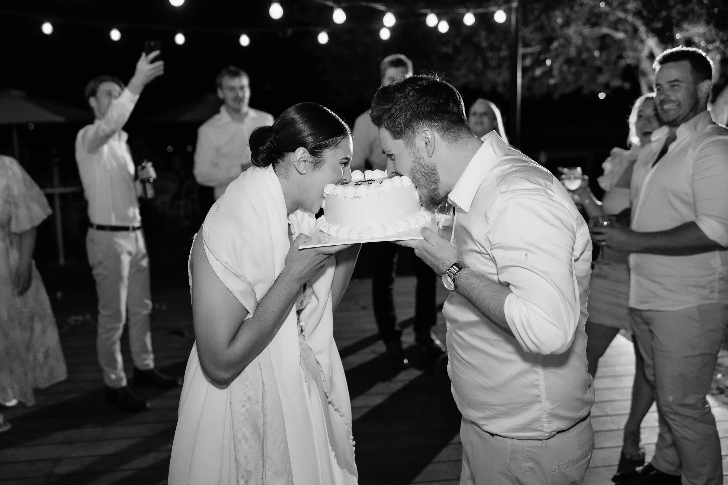 A couple is cutting their wedding cake with the bride and groom sharing a knife, surrounded by guests celebrating outdoors at night.