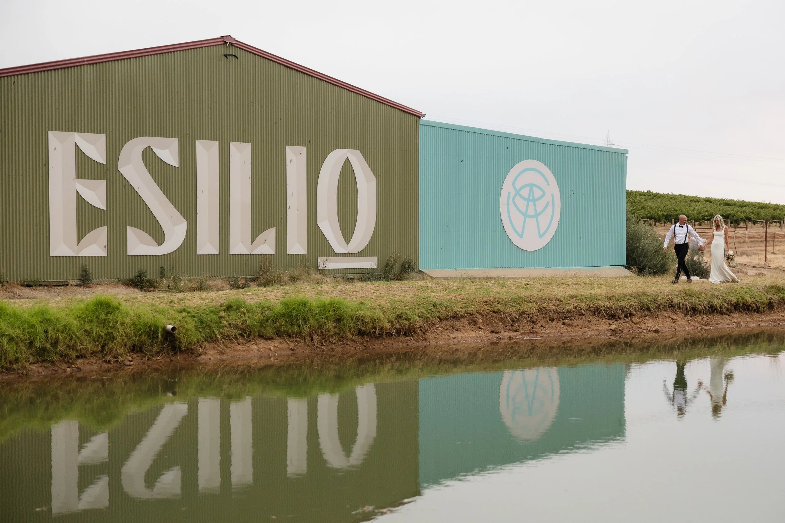 A couple in wedding attire walking hand in hand along a path near a body of water, with the venue name 'ESILO' displayed on a green industrial building and a logo on a blue section of the building.