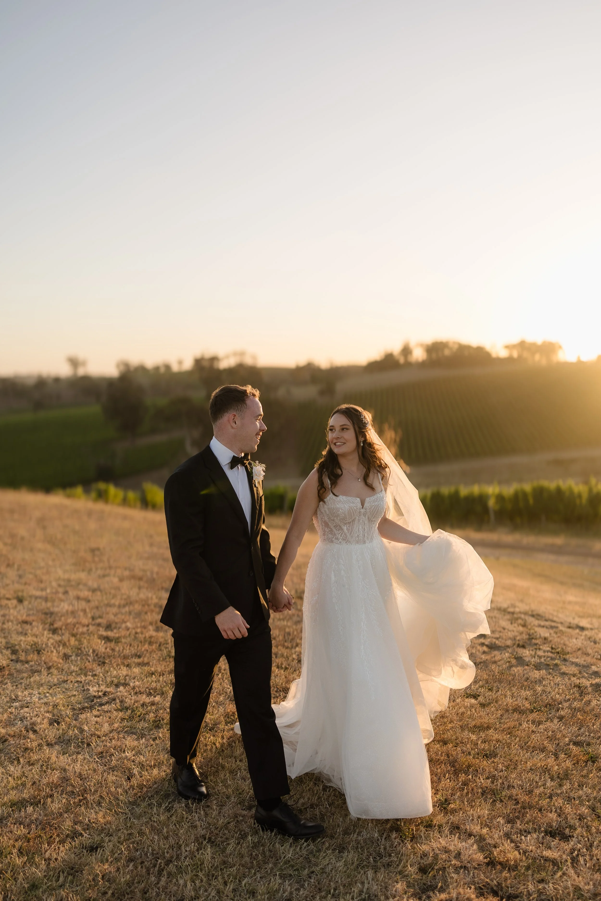 Bride and groom walking hand in hand through golden fields at sunset at Longview Vineyard, Macclesfield — Adelaide Hills vineyard wedding portrait with glowing backlight, bride in a flowing strapless lace gown with veil billowing in the breeze, groom