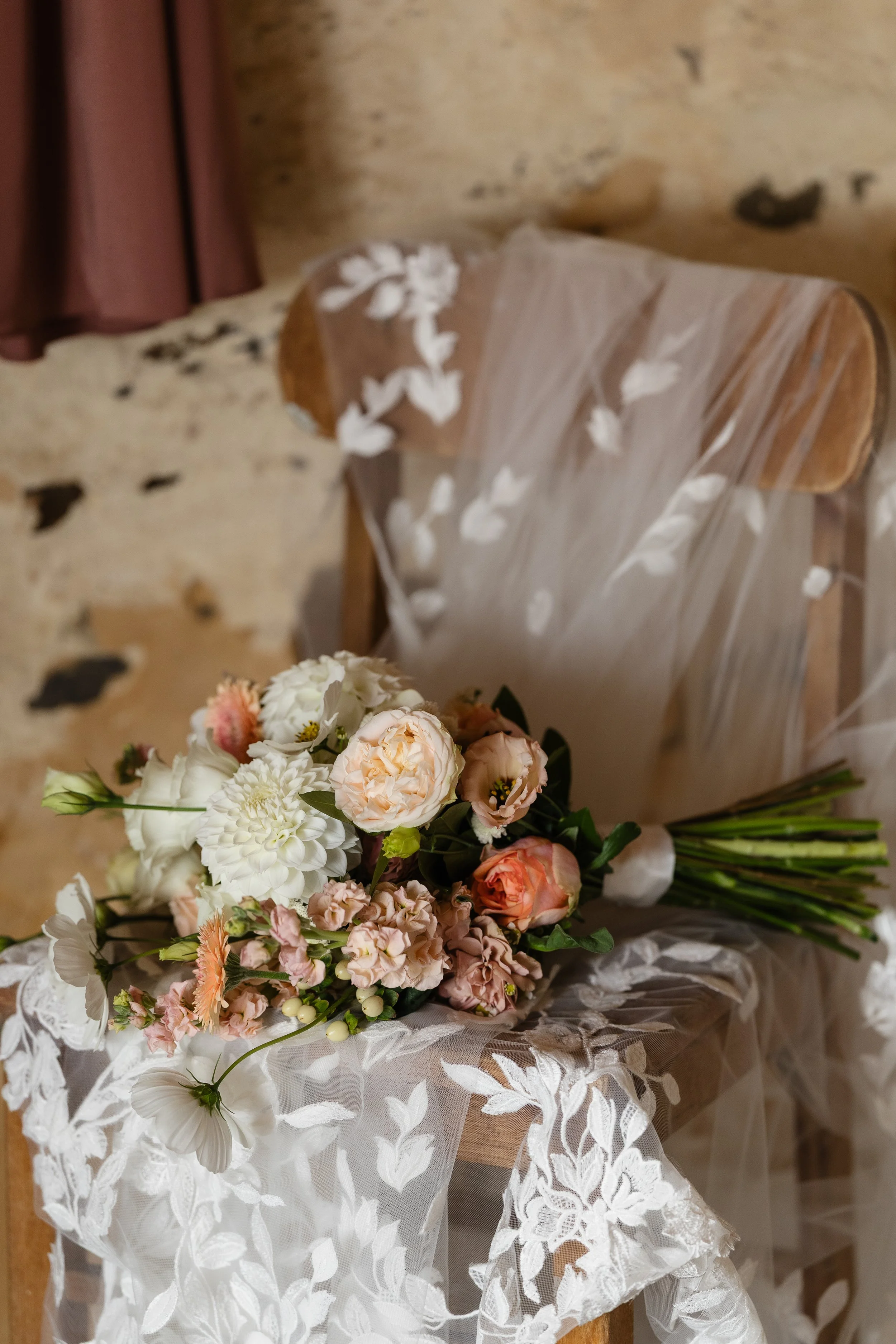 A bouquet of mixed white and peach-colored flowers resting on a wooden chair draped with a sheer, embroidered white cloth. The background features a rustic stone wall.