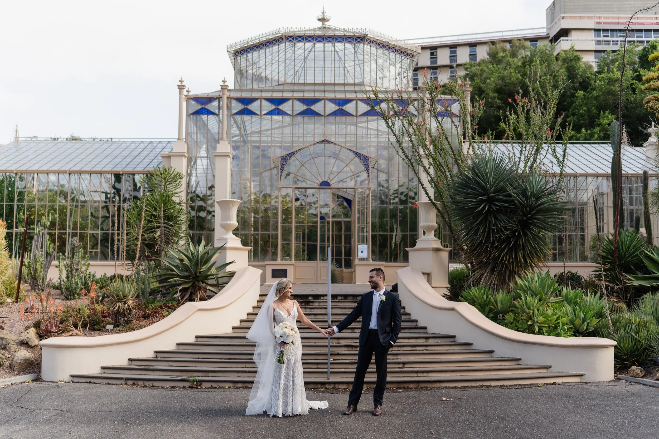 A bride and groom holding hands on a staircase outside a glass conservatory. The bride wears a white lace wedding dress with a bouquet, and the groom is in a dark suit with a boutonniere. The conservatory has large glass panels with a decorative roof, surrounded by desert plants and greenery.