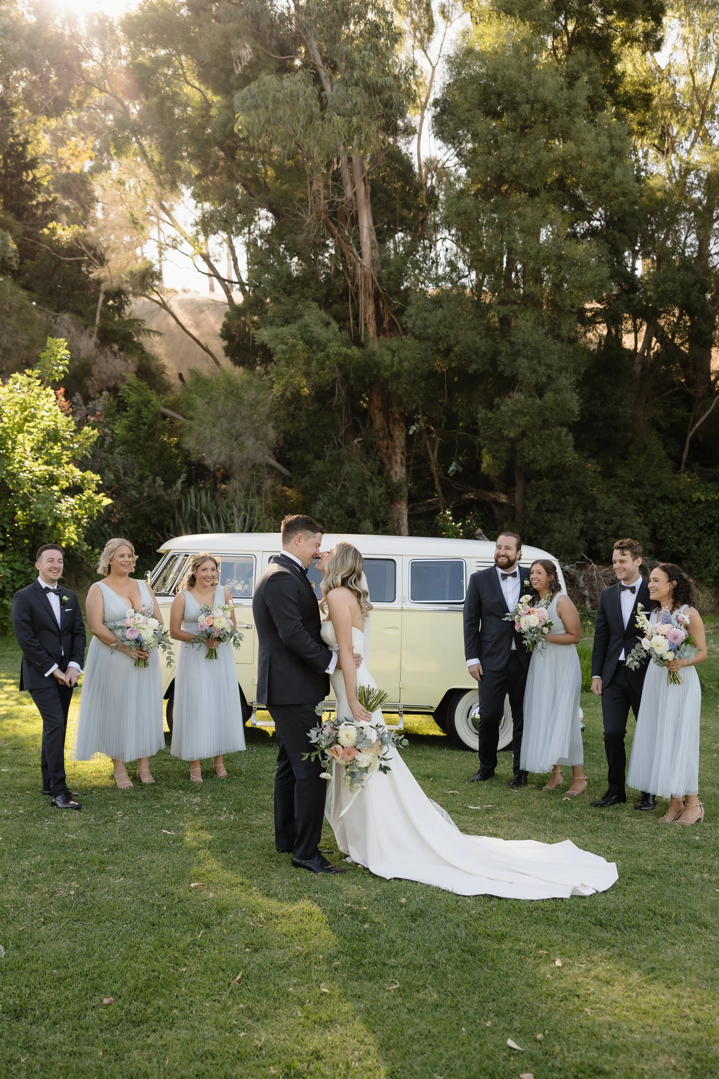 Bride and groom sharing a laugh with their wedding party in front of a vintage cream Volkswagen kombi van at Netherhill Farm, Adelaide Hills — bridesmaids in soft sage tulle dresses holding pink and white bouquets, groomsmen in black tuxedos on a sun