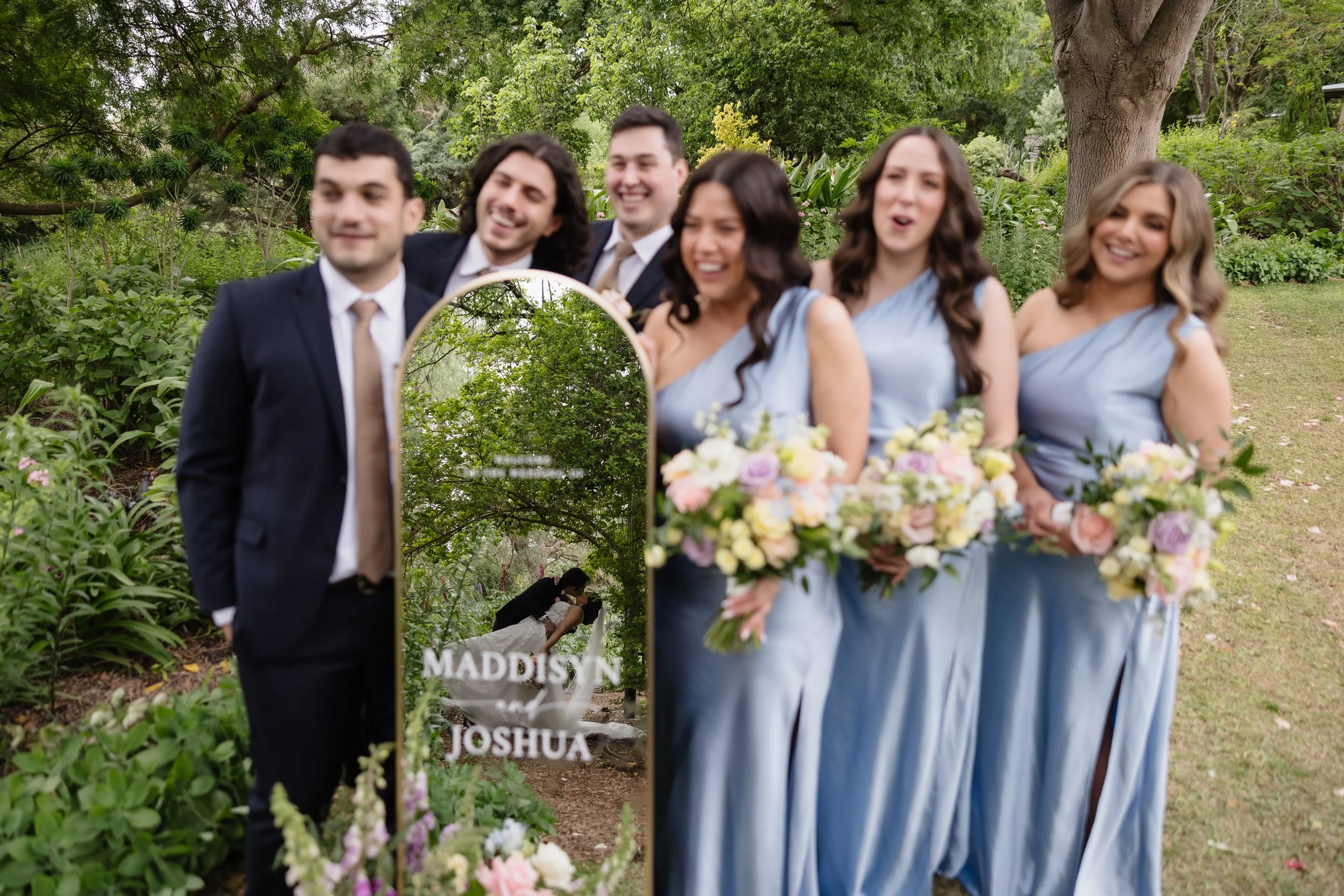 Wedding party standing outdoors with wedding mirror reflecting a couple kissing, wedding couple image digitally added on mirror with names Maddisyn and Joshua.