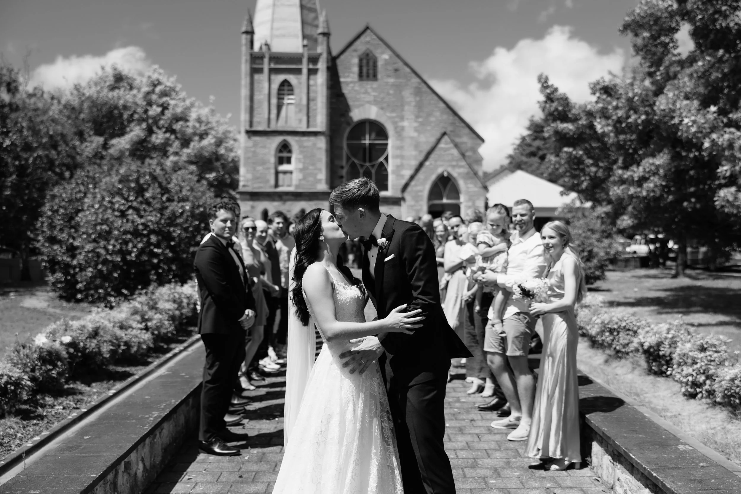 Bride and groom sharing a kiss outside a historic stone church in Hahndorf, Adelaide Hills, surrounded by cheering guests — black and white wedding photo, bride in lace gown with cathedral veil, groom in black tuxedo