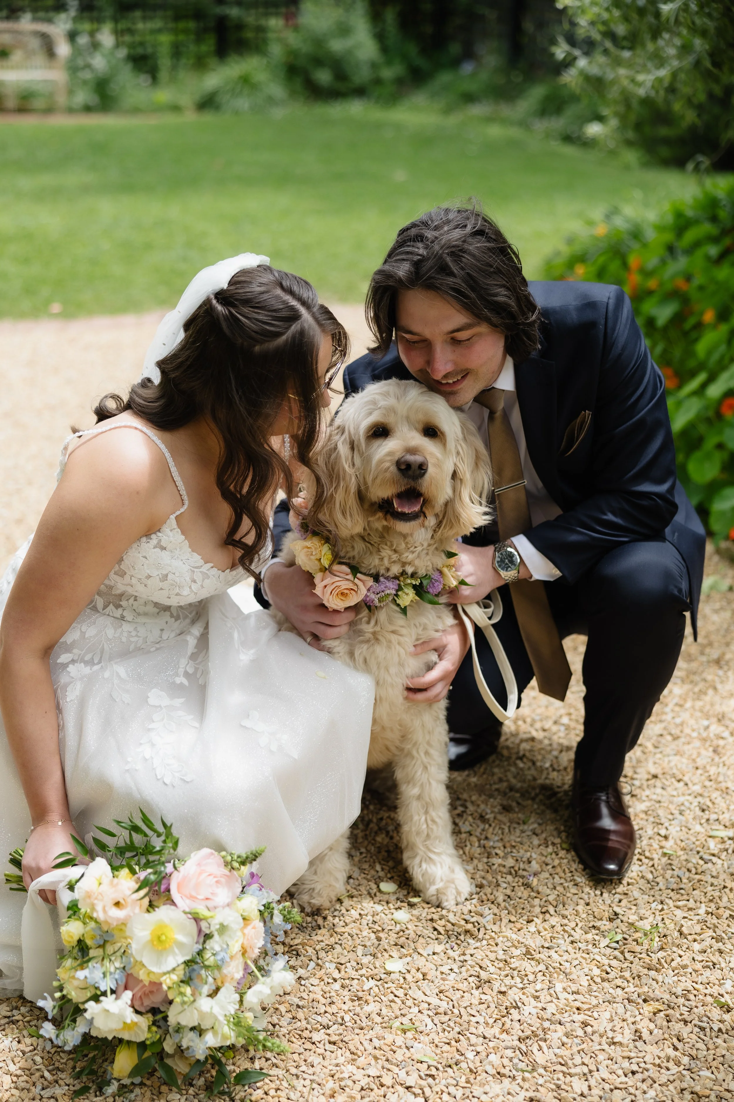A bride and groom kneel down, smiling, with a golden retriever dog wearing a floral collar between them. The bride is holding a bouquet of flowers, and the photo is taken outdoors in a garden setting.