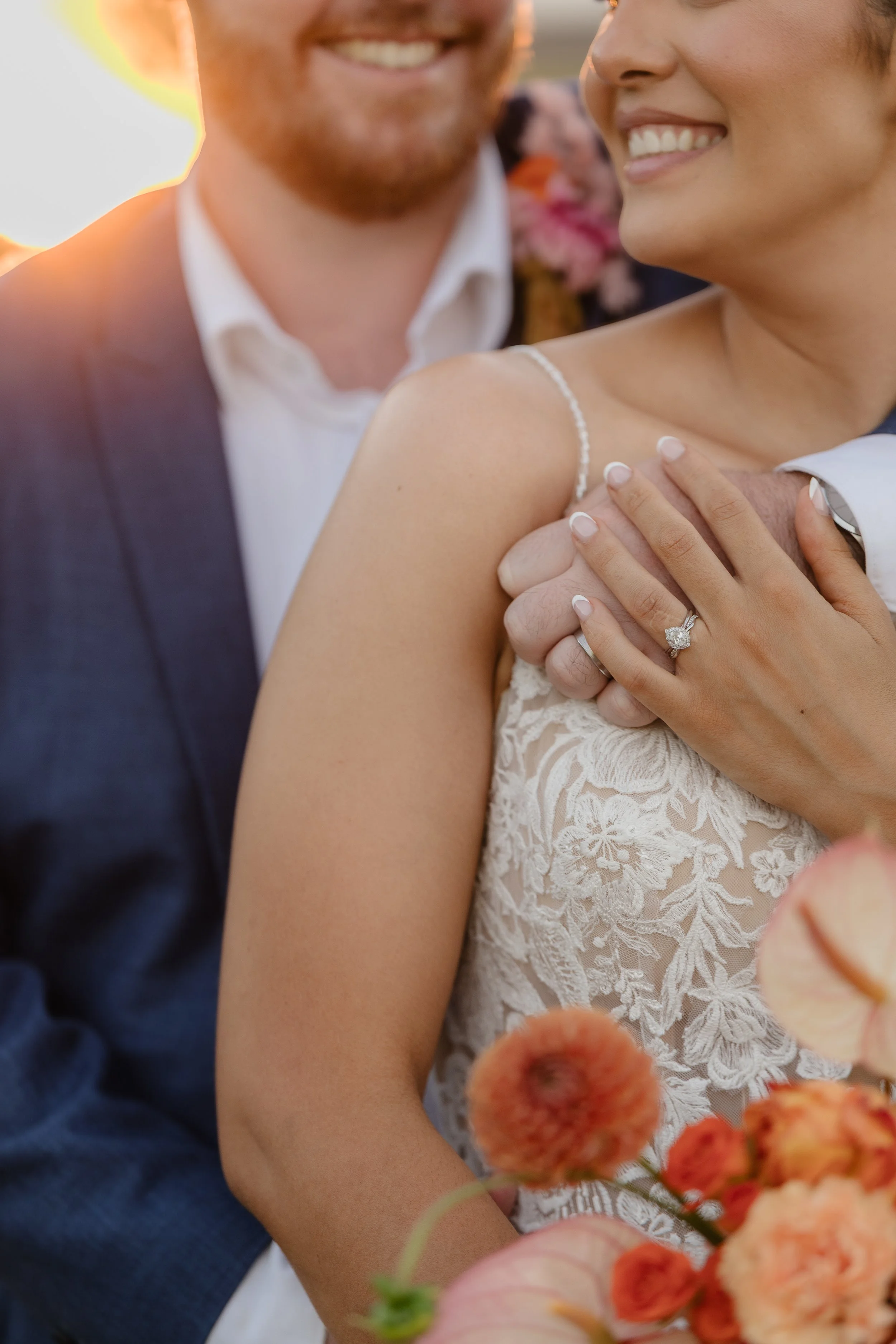 A close-up of a smiling bride and groom at their wedding, with the bride wearing a lace wedding dress and the groom in a dark suit. The bride's hand, showing her engagement ring, rests on the groom's arm, and they are surrounded by flowers.