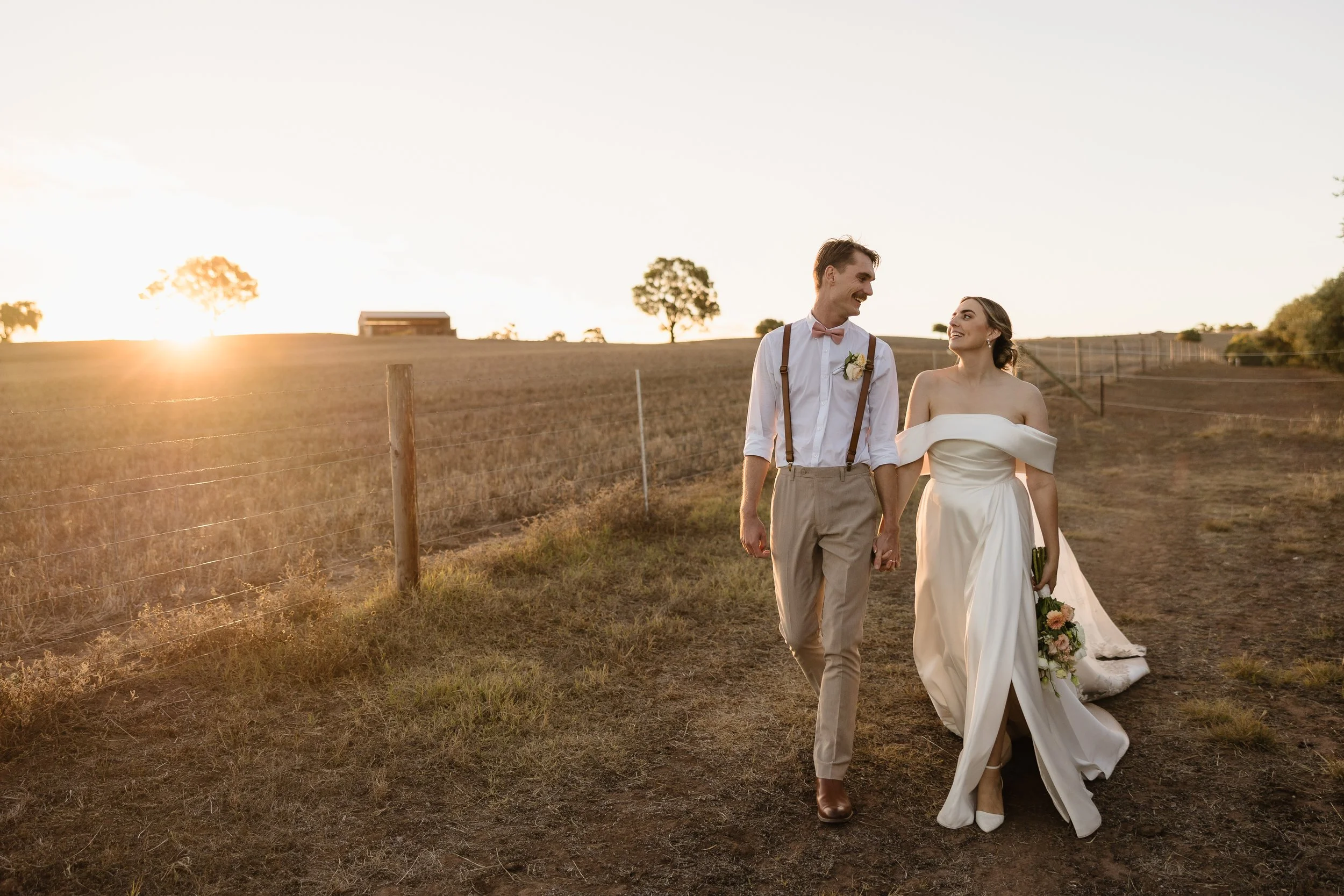 A newlywed couple walking hand in hand on a dirt path in a rural landscape at sunset. The groom wears a white shirt with rolled-up sleeves, beige pants, suspenders, a pink bowtie, and a boutonniere. The bride wears a white off-the-shoulder wedding gown with a train and holds a bouquet of flowers. They smile at each other as they walk.