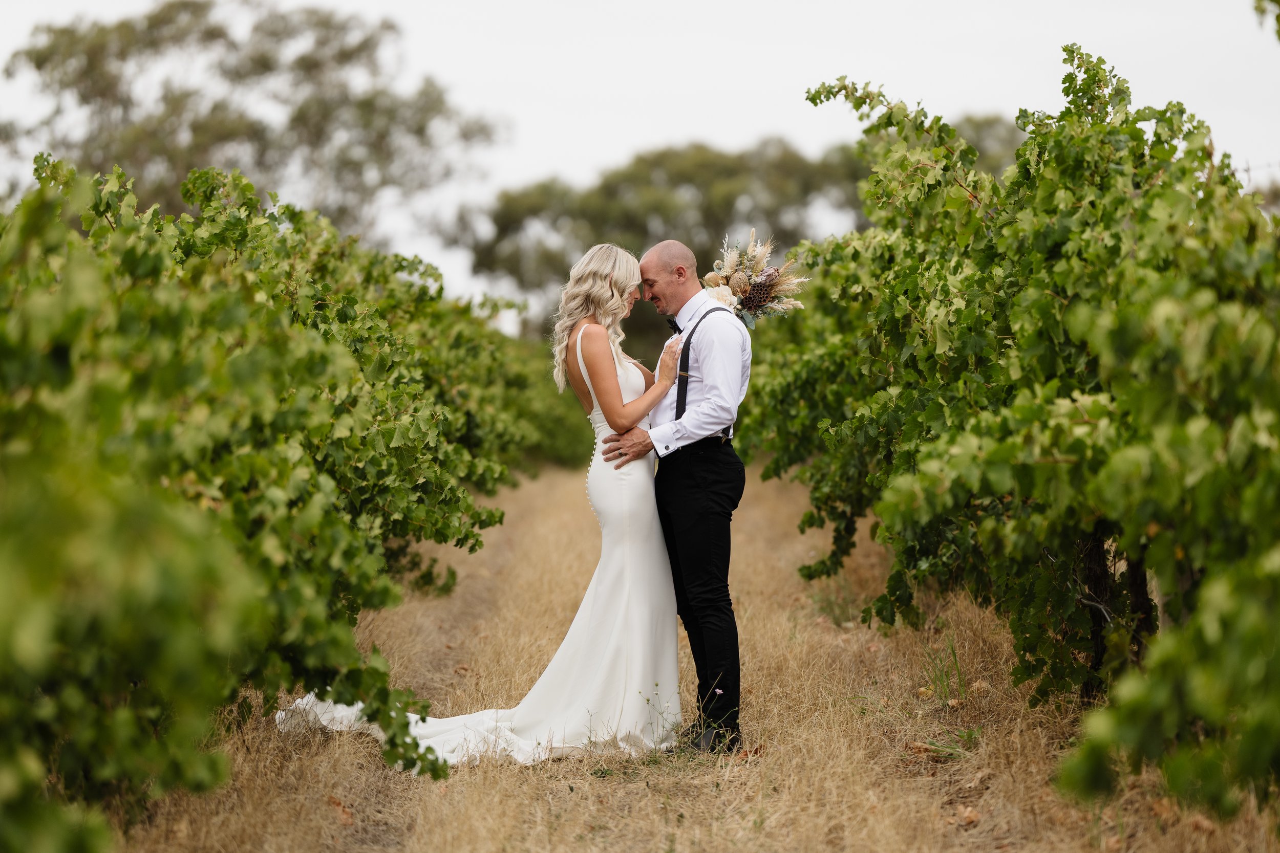 A bride and groom in wedding attire standing close together in a vineyard, touching foreheads, with a bouquet of flowers on the groom's shoulder, surrounded by green grapevines.