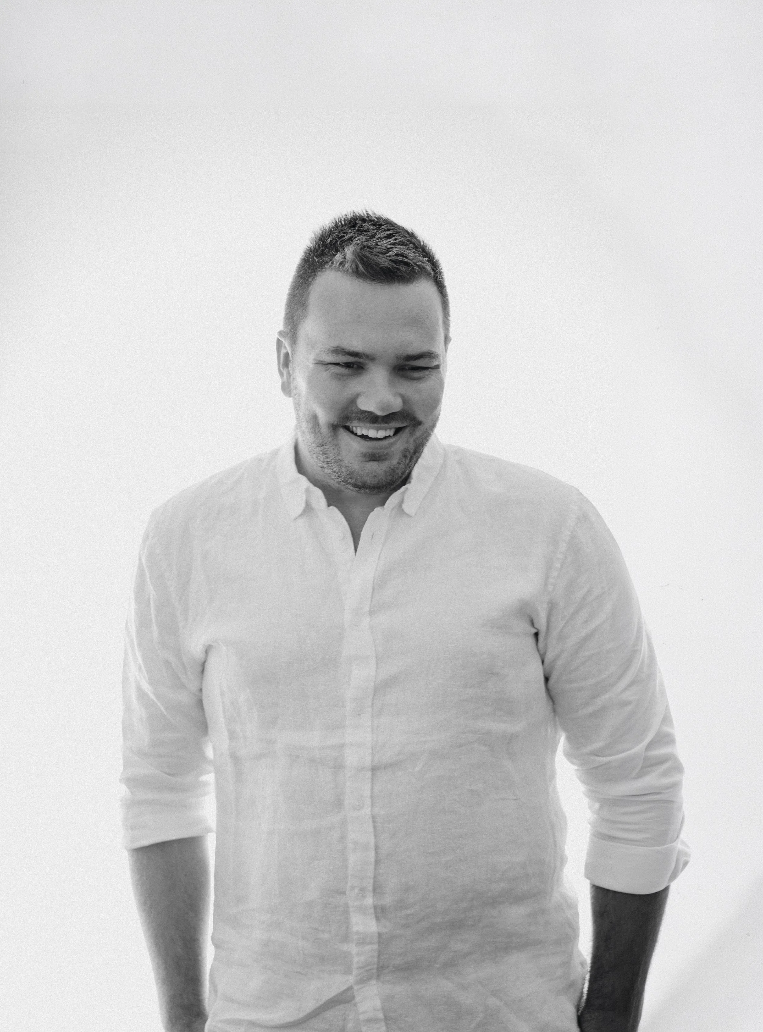 A black and white photo of a smiling man with short hair, wearing a white button-up shirt with rolled-up sleeves, standing against a plain light background.