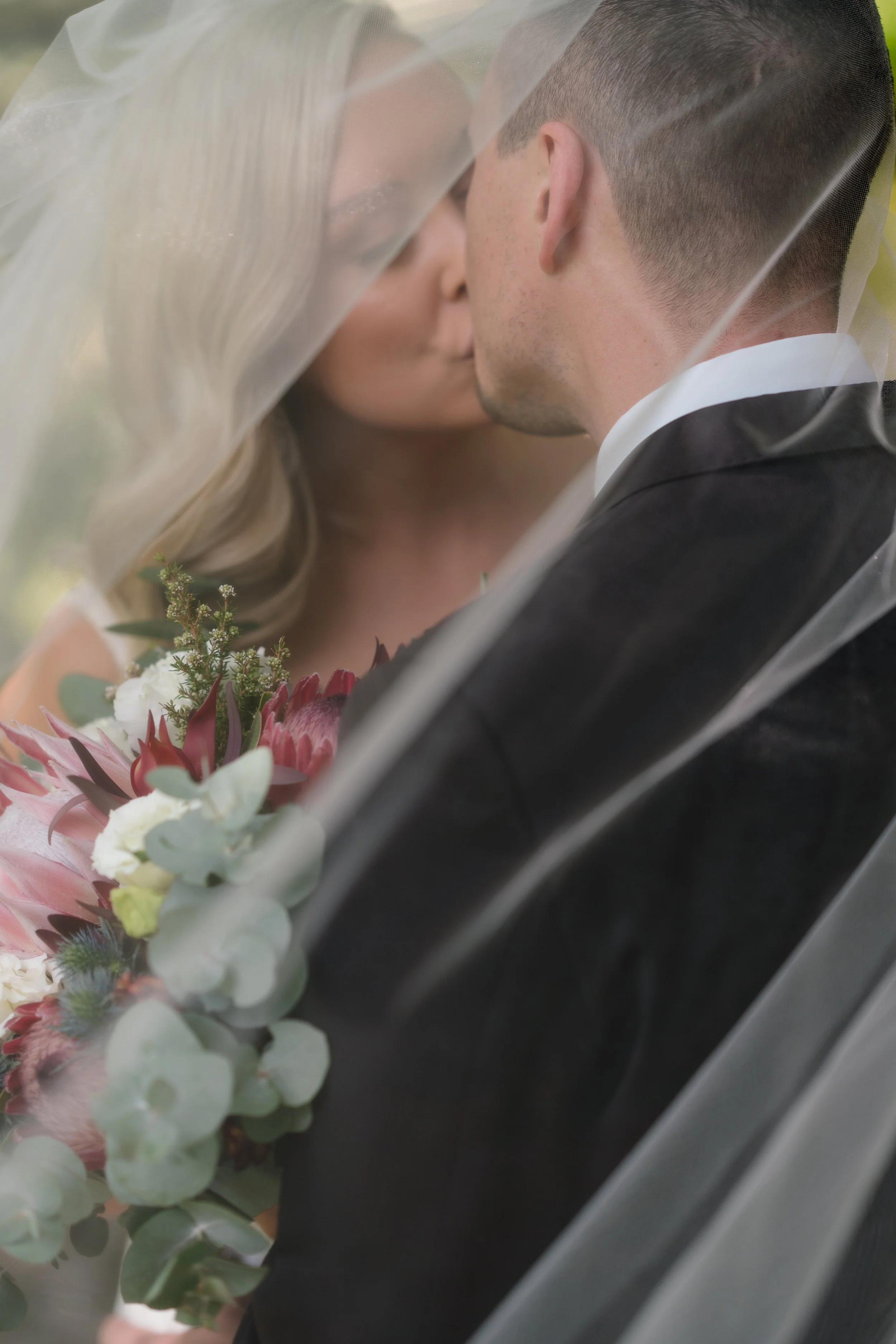 A bride and groom kiss under a sheer veil, with the bride holding a bouquet of pink and white flowers.