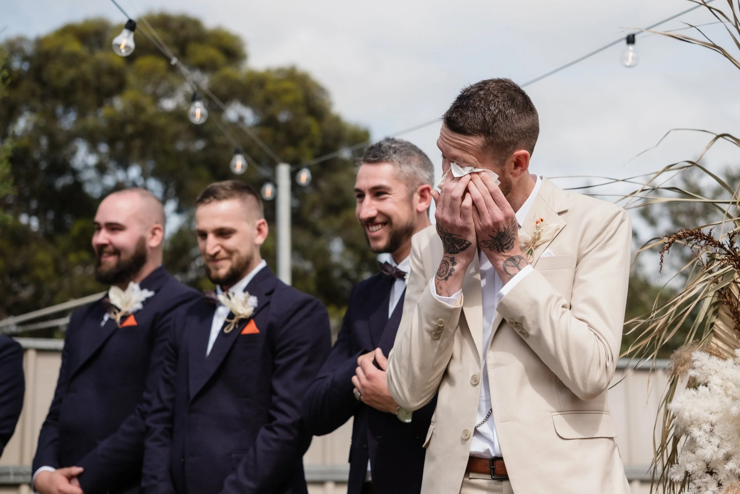 Emotional groom crying happy tears during the bridal entrance at an outdoor backyard wedding in Adelaide — groom in a cream suit with groomsmen in navy, pampas grass and dried floral arch, festoon lights overhead