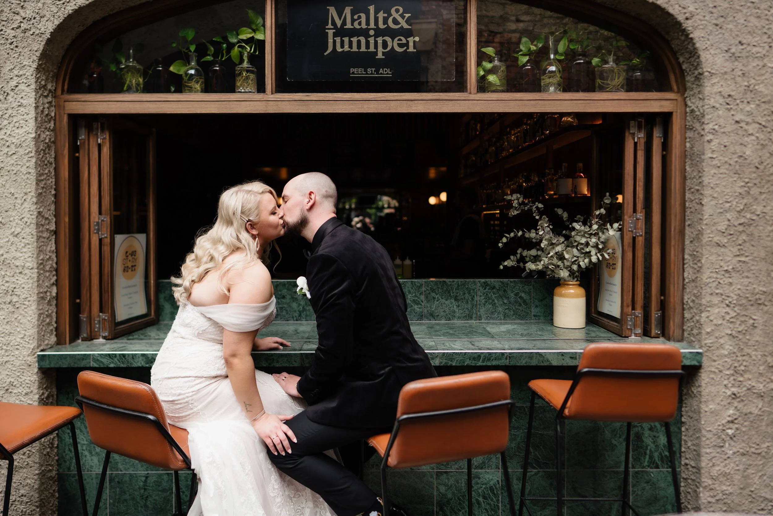 A couple in wedding attire sharing a kiss at a small bar or cafe with green marble countertop and brown chairs.