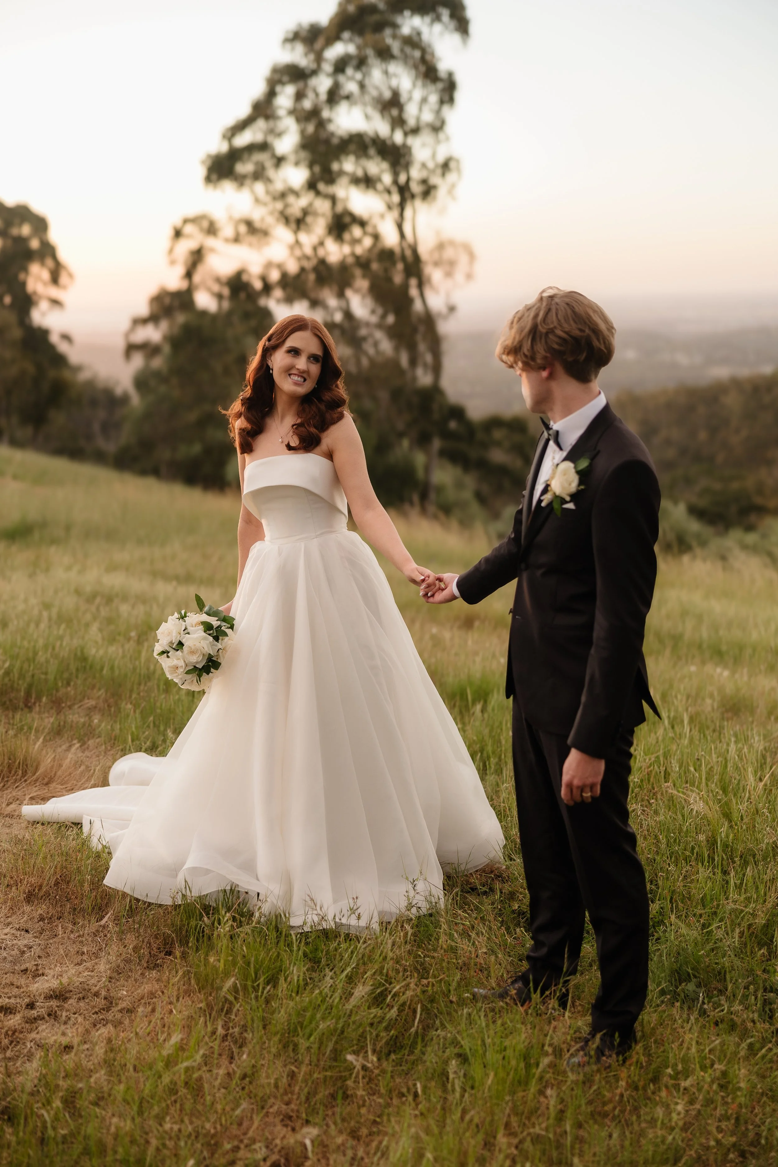 Bride and groom holding hands in golden hour light on the rolling green hills of Glen Ewin Estate, Adelaide Hills — bride wearing a strapless tulle ballgown with white rose bouquet, groom in black tuxedo