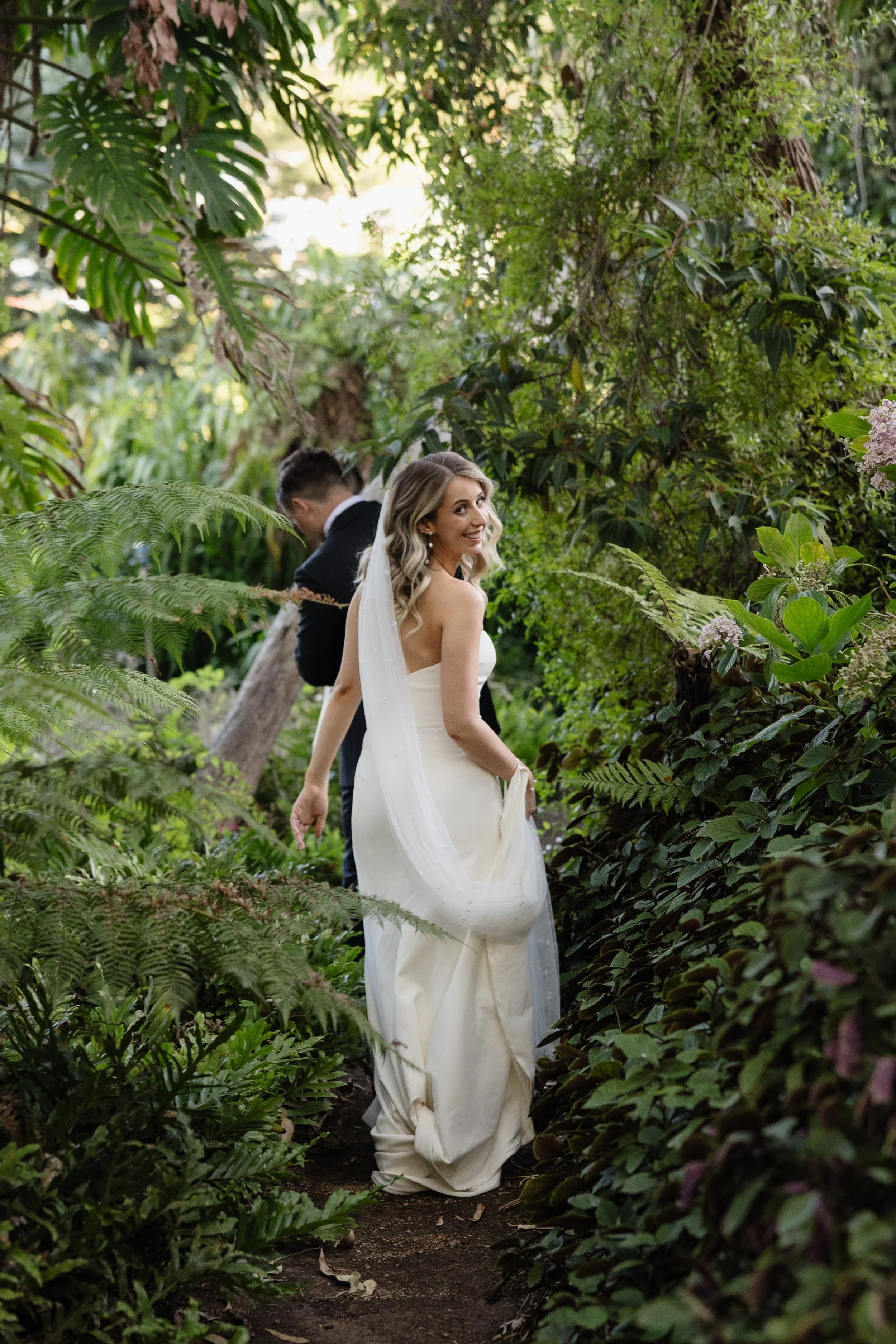 Bride glancing back over her shoulder while walking through a lush fern-lined garden path at Netherhill Farm, Adelaide Hills — bride in a sleek strapless white gown with veil, groom in black suit following behind through tropical greenery