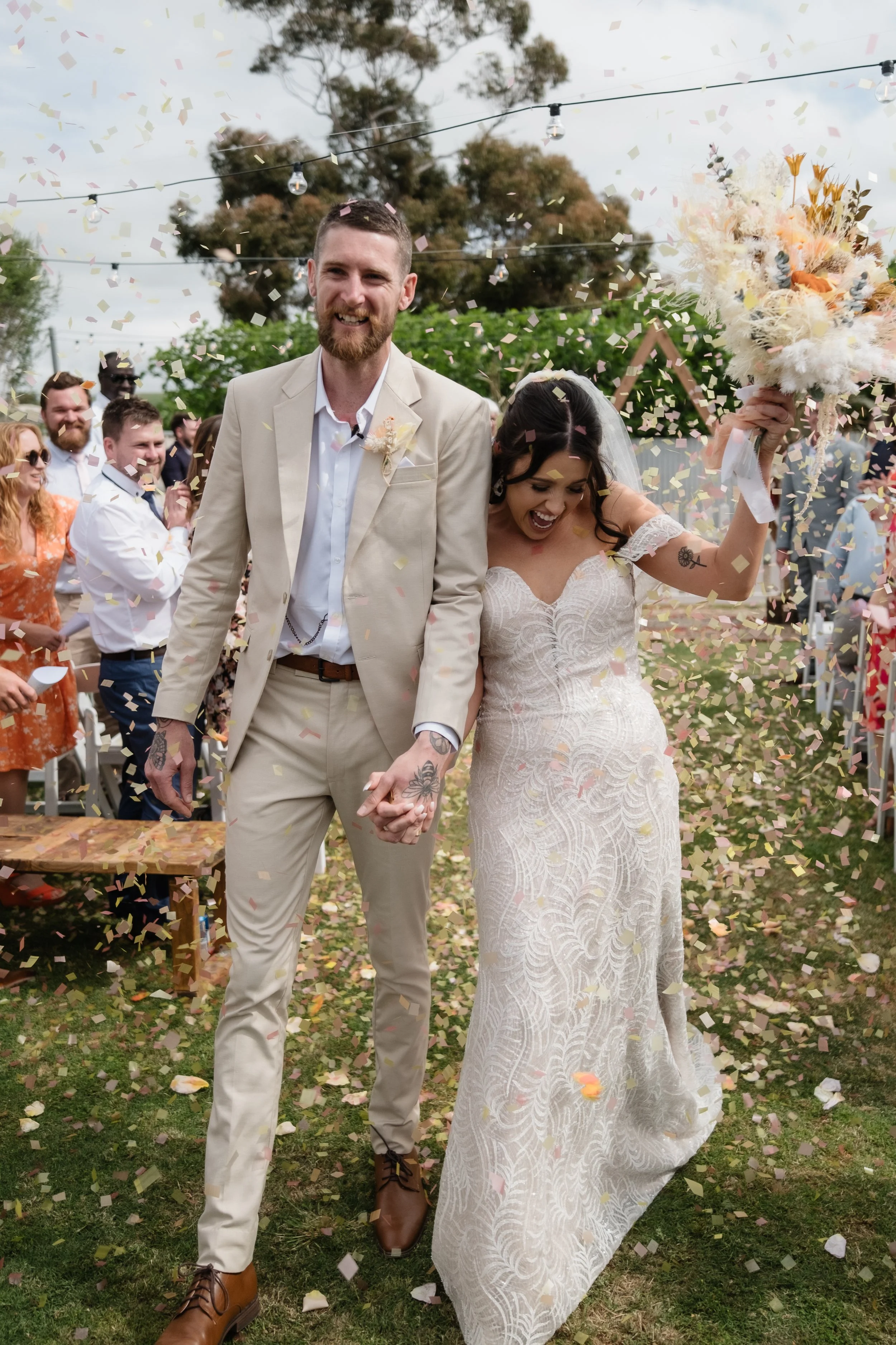Bride and groom walking back down the aisle in a shower of colourful confetti at a relaxed backyard wedding in Adelaide — groom in a tan suit, bride in a fitted lace gown holding a boho dried flower bouquet, guests cheering under festoon lights