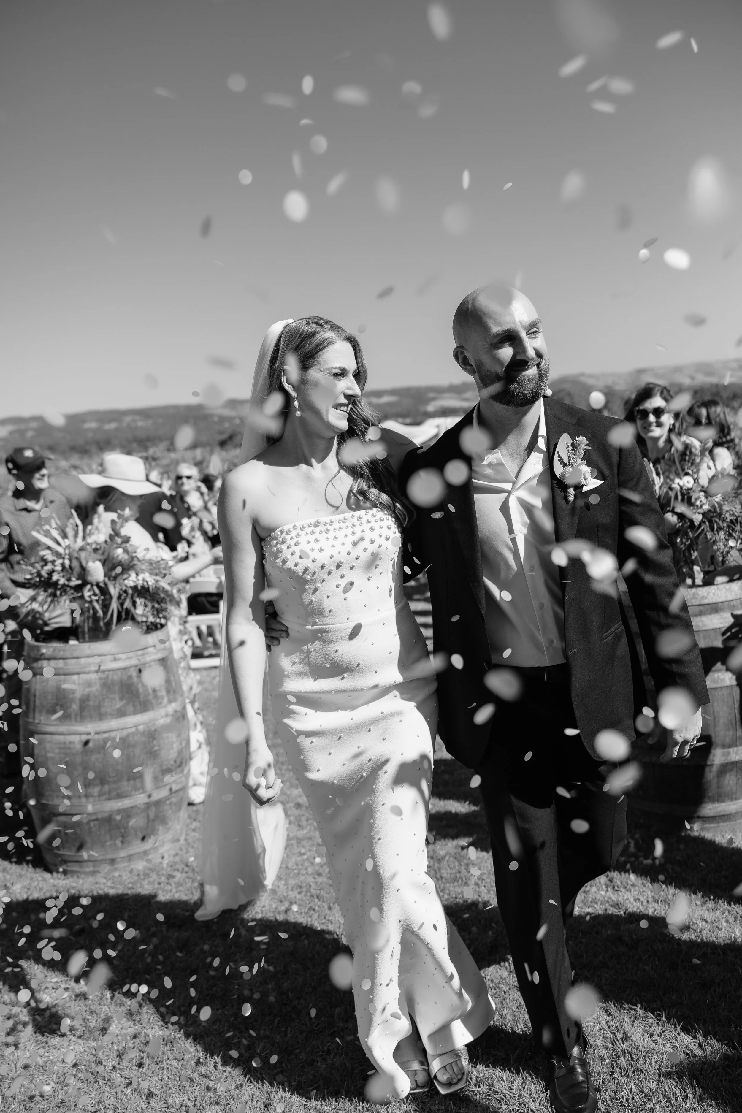 A bride and groom walking outdoors at their wedding, surrounded by guests and falling confetti, with scenery in the background.