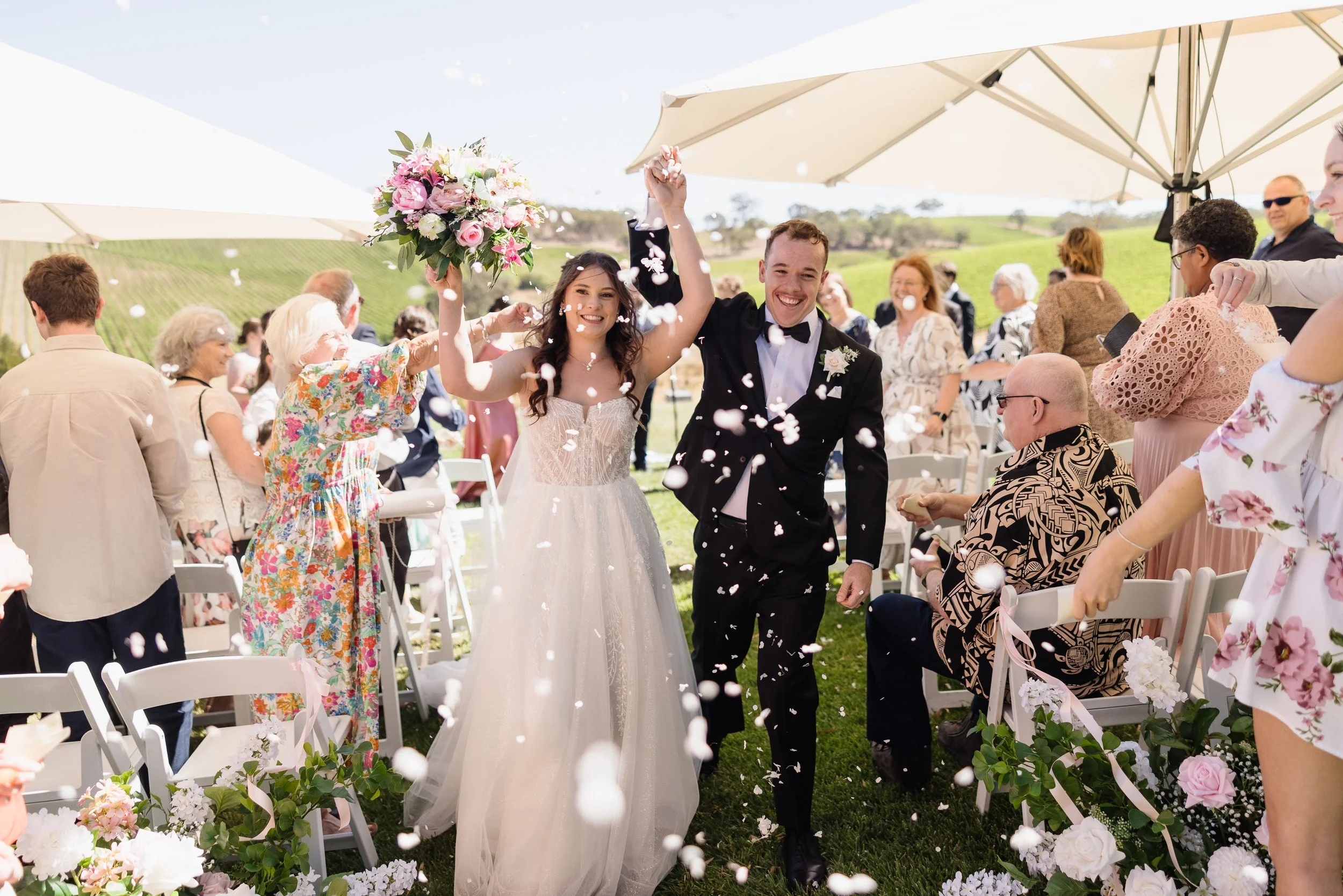 Bride and groom celebrating their recessional in a shower of white confetti at Longview Vineyard, Macclesfield — outdoor vineyard wedding ceremony with rolling green hills, bride in a strapless tulle ballgown holding a pink peony bouquet, groom in bl