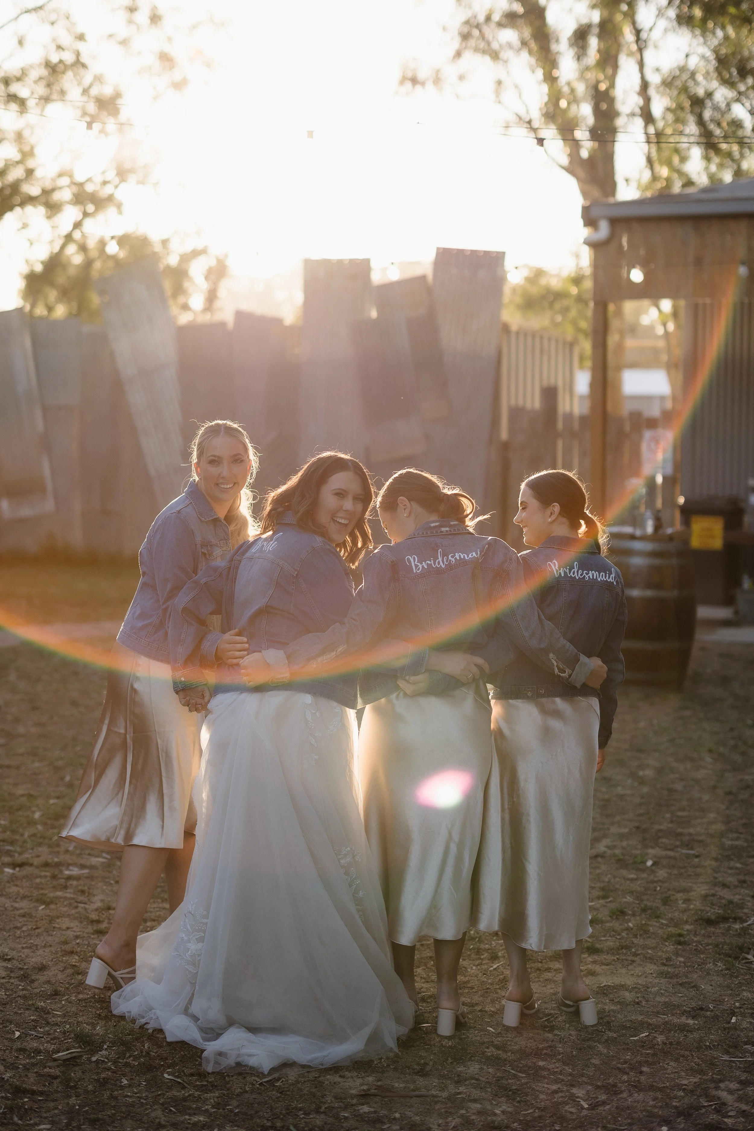 Four women, with some wearing bridal and bridesmaid jackets, enjoying time outdoors during sunset, with a rustic wooden fence in the background.