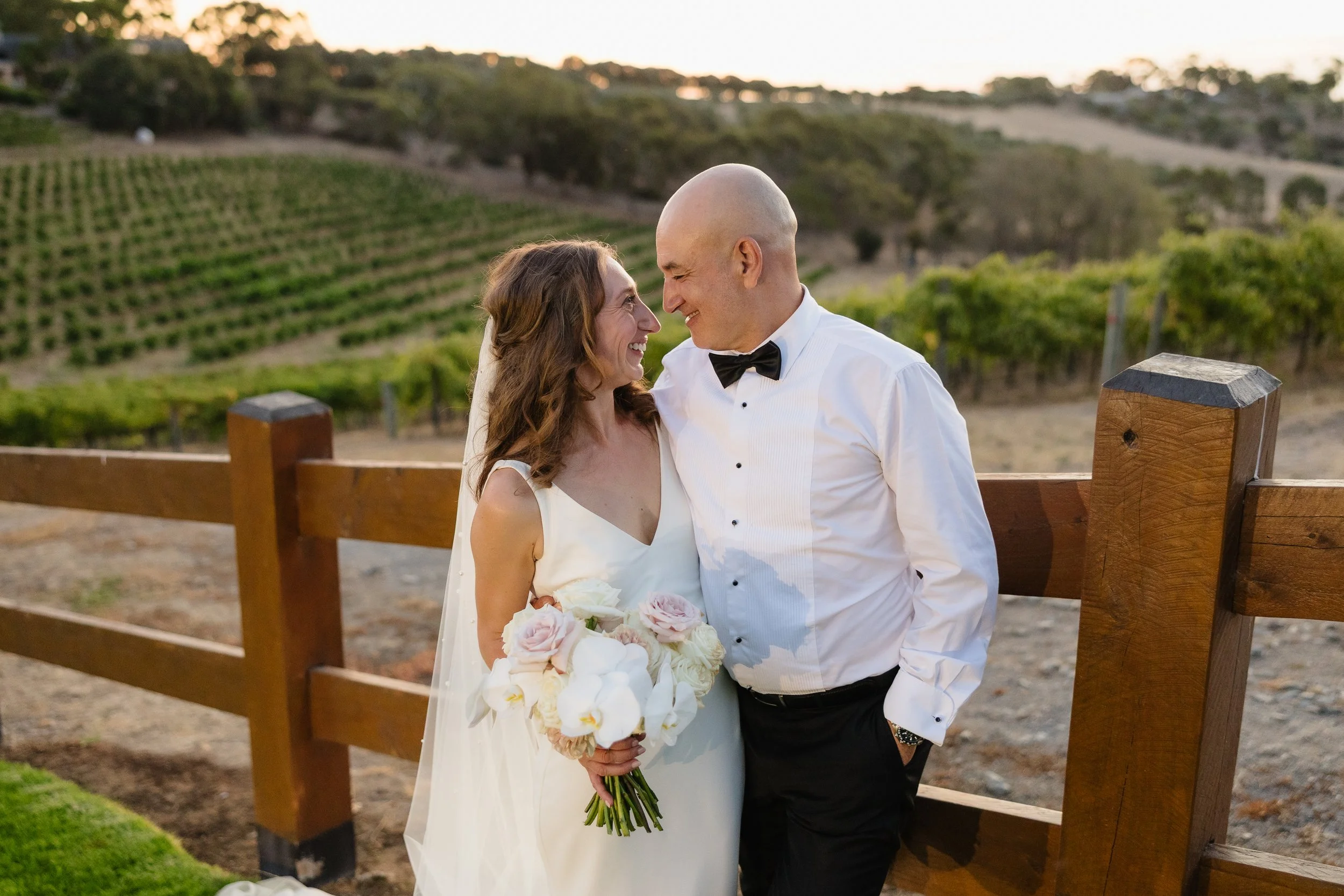 A newlywed couple smiling and looking into each other's eyes on their wedding day, standing outdoors with a vineyard in the background at sunset. The bride holds a bouquet of white and pink roses, and the groom is dressed in a white tuxedo shirt with a black bow tie and black pants.