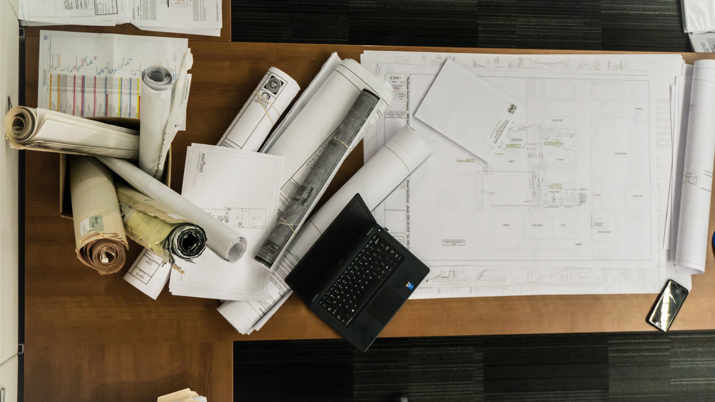 An office desk with architectural blueprints, rolled-up drawings, a laptop, and a cell phone.
