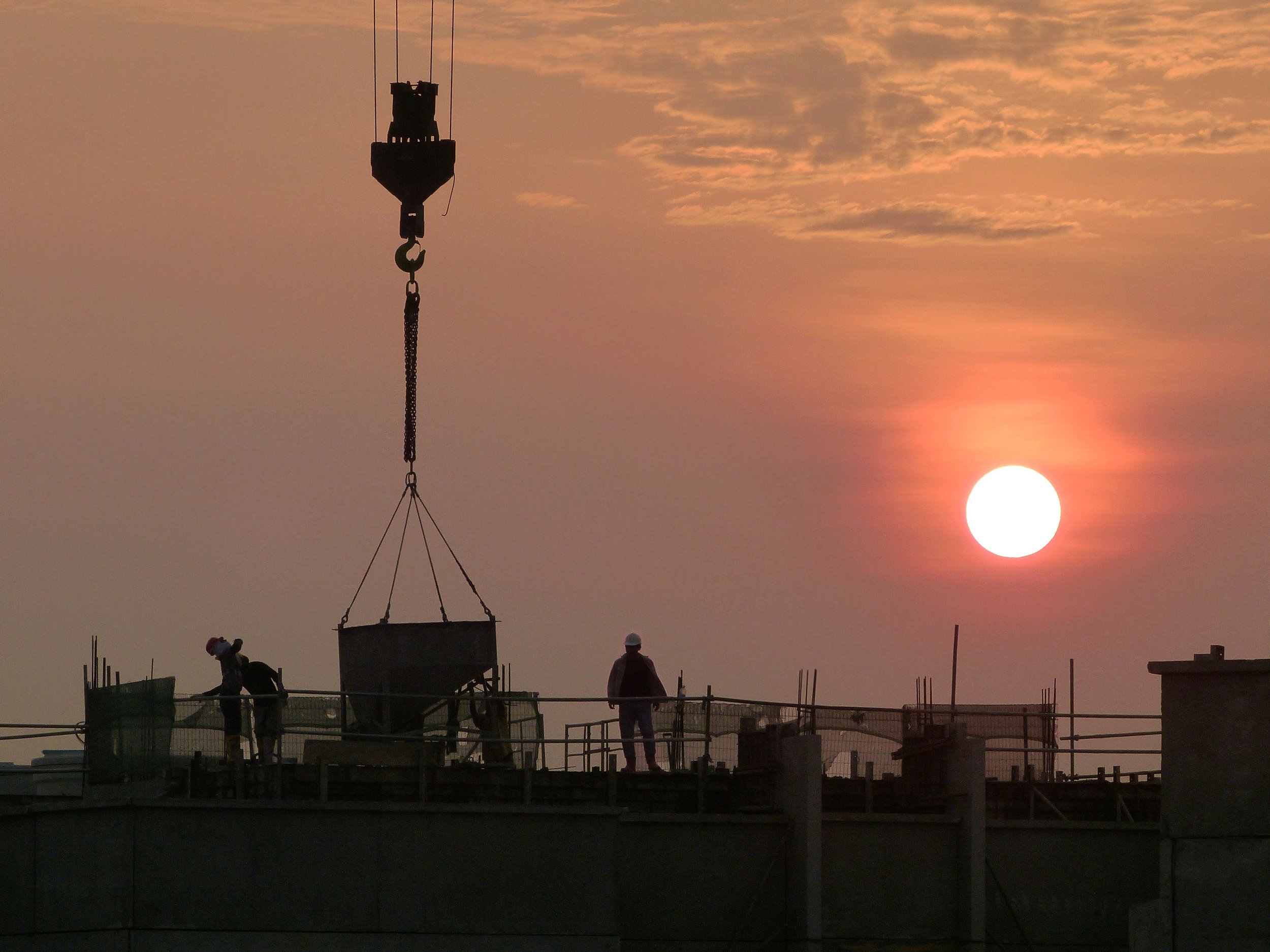 Construction workers on a building site at sunset, with a crane lifting materials and the sun low in the sky.