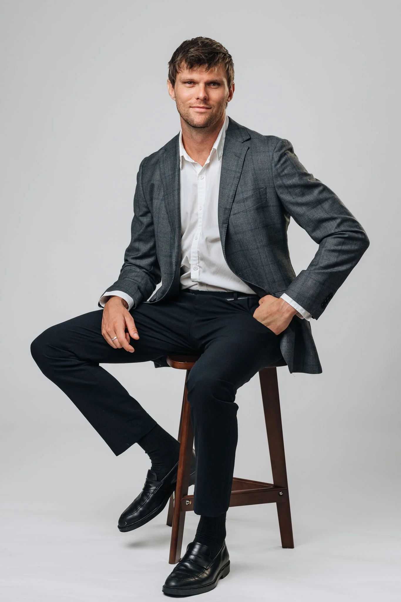 A man in a gray suit with a white dress shirt, sitting on a wooden stool against a plain white background.