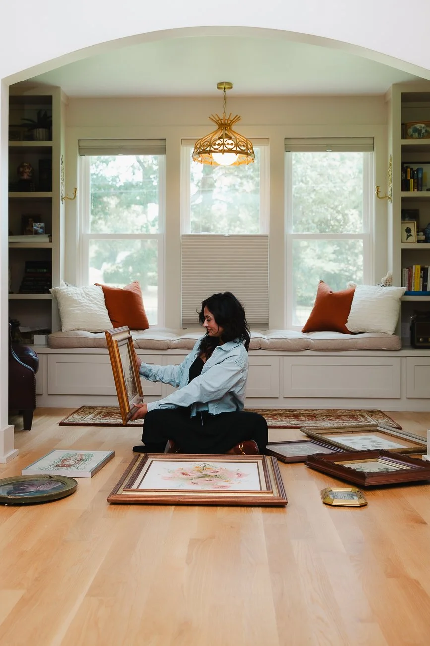A woman sorting framed artwork on a hardwood floor in a bright living room with large windows and built-in window seat.