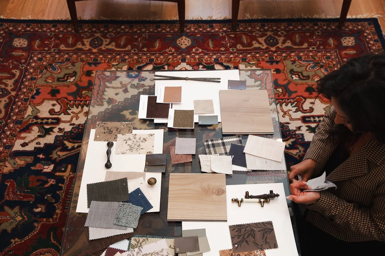 Interior design consultation with fabric samples, paint swatches, and wood finishes laid out on a glass table over an ornate red and blue patterned rug.
