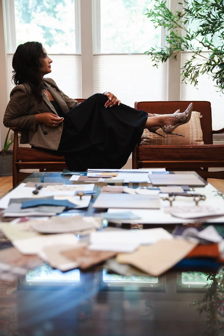 A woman with dark hair, wearing a checkered blazer and black dress, sitting on a sofa with her legs crossed and feet resting on a cushion. She appears to be in an office or lounge with large windows, a table with various papers and objects in front, and a large green plant nearby.